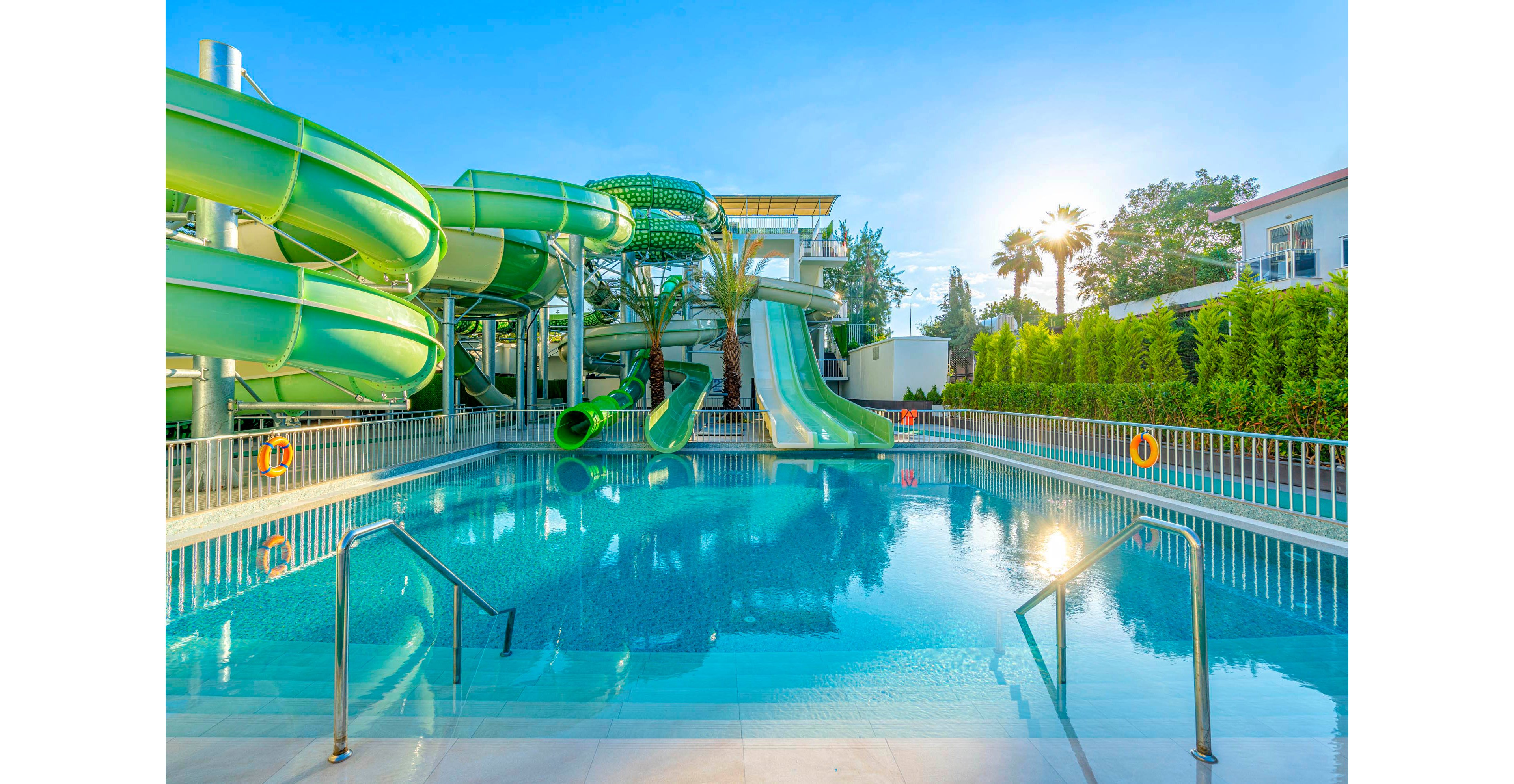 A large outdoor swimming pool is featured in the foreground, with multiple green water slides curving above and leading into the pool. The setting is a modern water park or resort, surrounded by palm trees, greenery, and nearby buildings. The scene is brightly lit by the morning sun, creating a vibrant and inviting atmosphere.