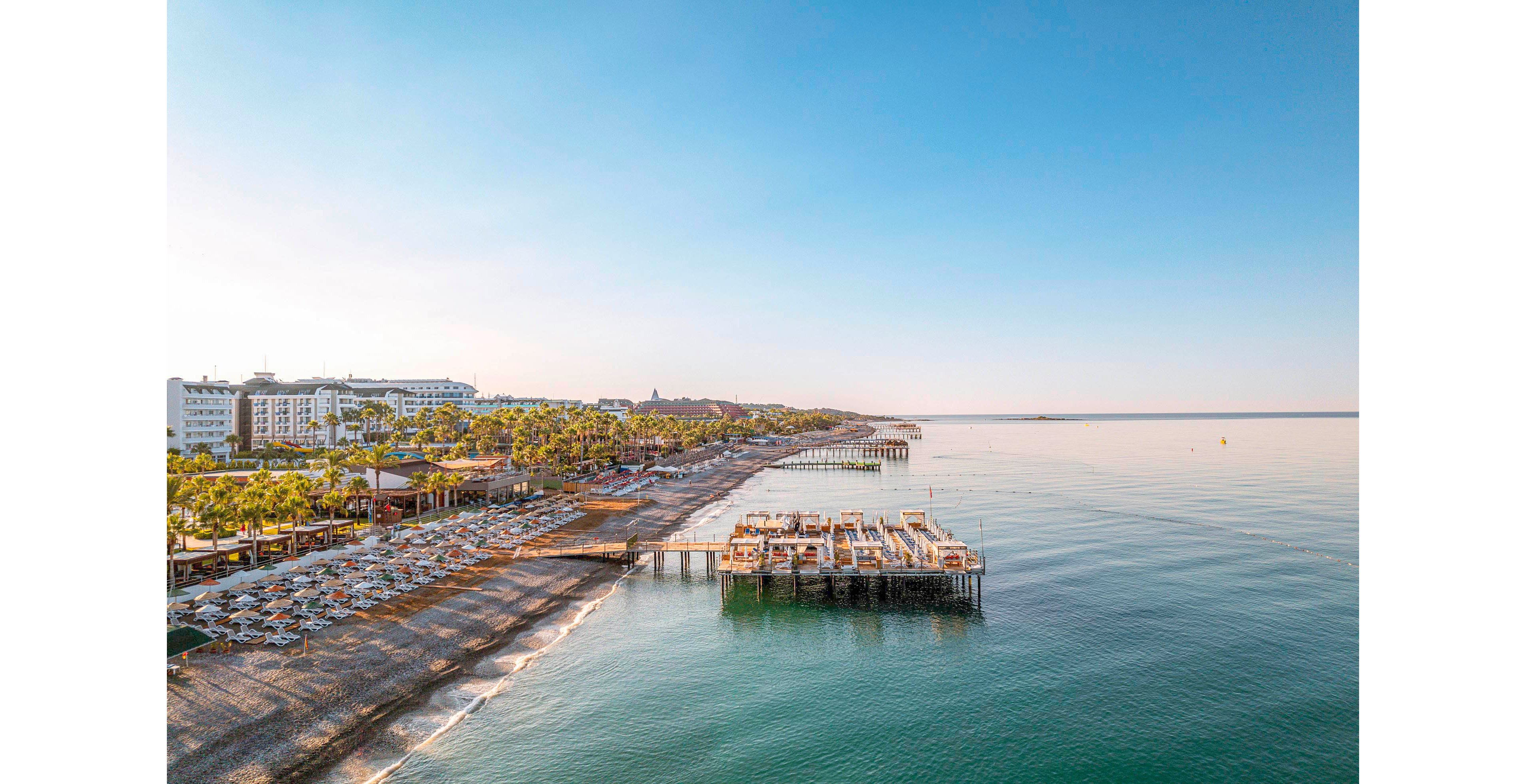 The image shows a vibrant beachfront resort with rows of sun loungers and umbrellas along the sandy shore. Multiple wooden piers extend into the calm, clear sea, creating inviting spaces for relaxation. The setting is tranquil, with palm trees and resort buildings visible in the background under a bright blue sky. The overall mood is peaceful and leisurely, ideal for vacation or holiday activities.