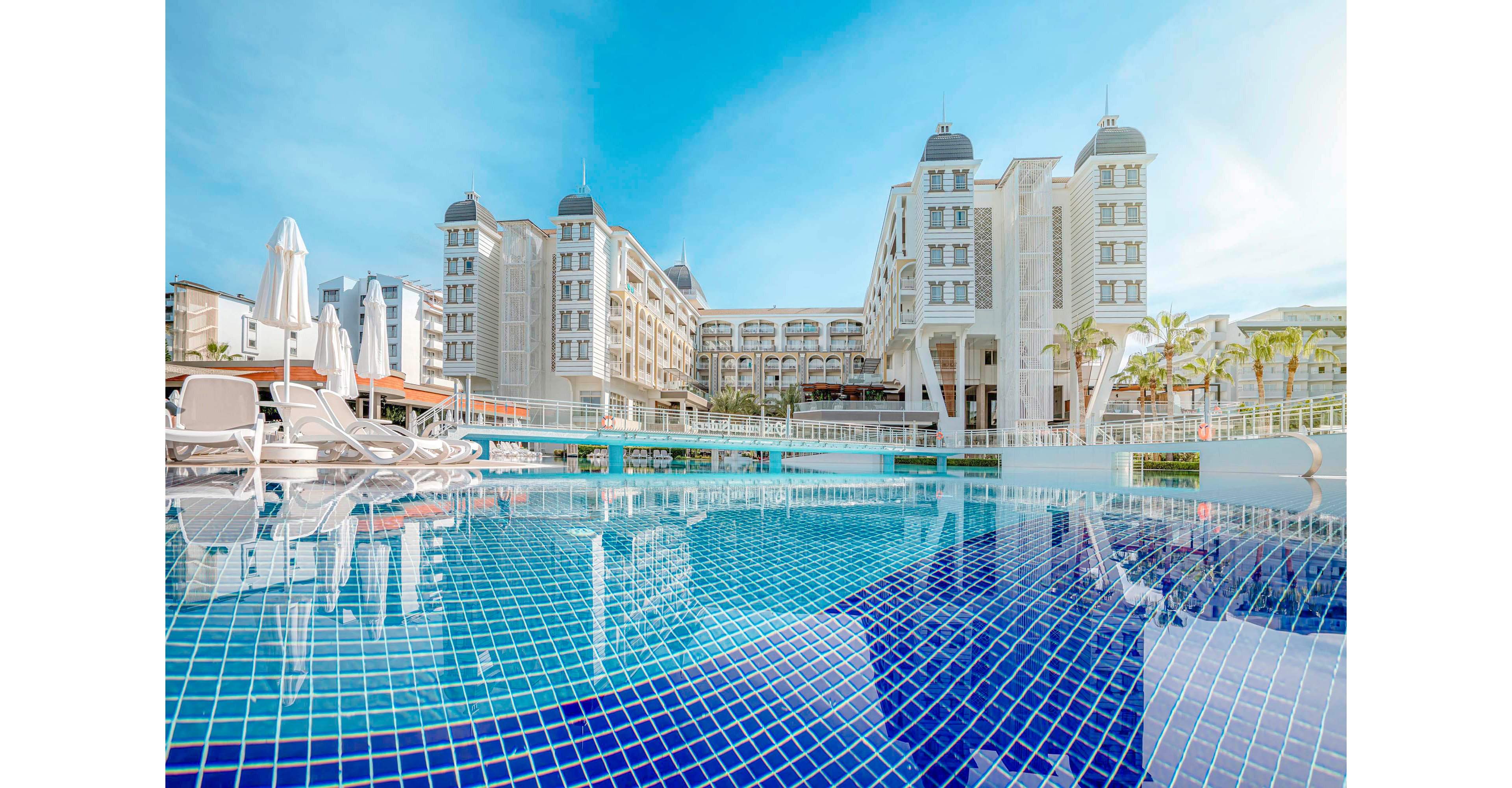 A grand hotel building stands behind a spacious outdoor swimming pool with blue mosaic tiles. The scene is set on a sunny day, with clear skies and reflections of the architecture in the water. White lounge chairs and umbrellas line the poolside, suggesting a relaxing and upscale atmosphere. No visible text or numbers are present in the image.