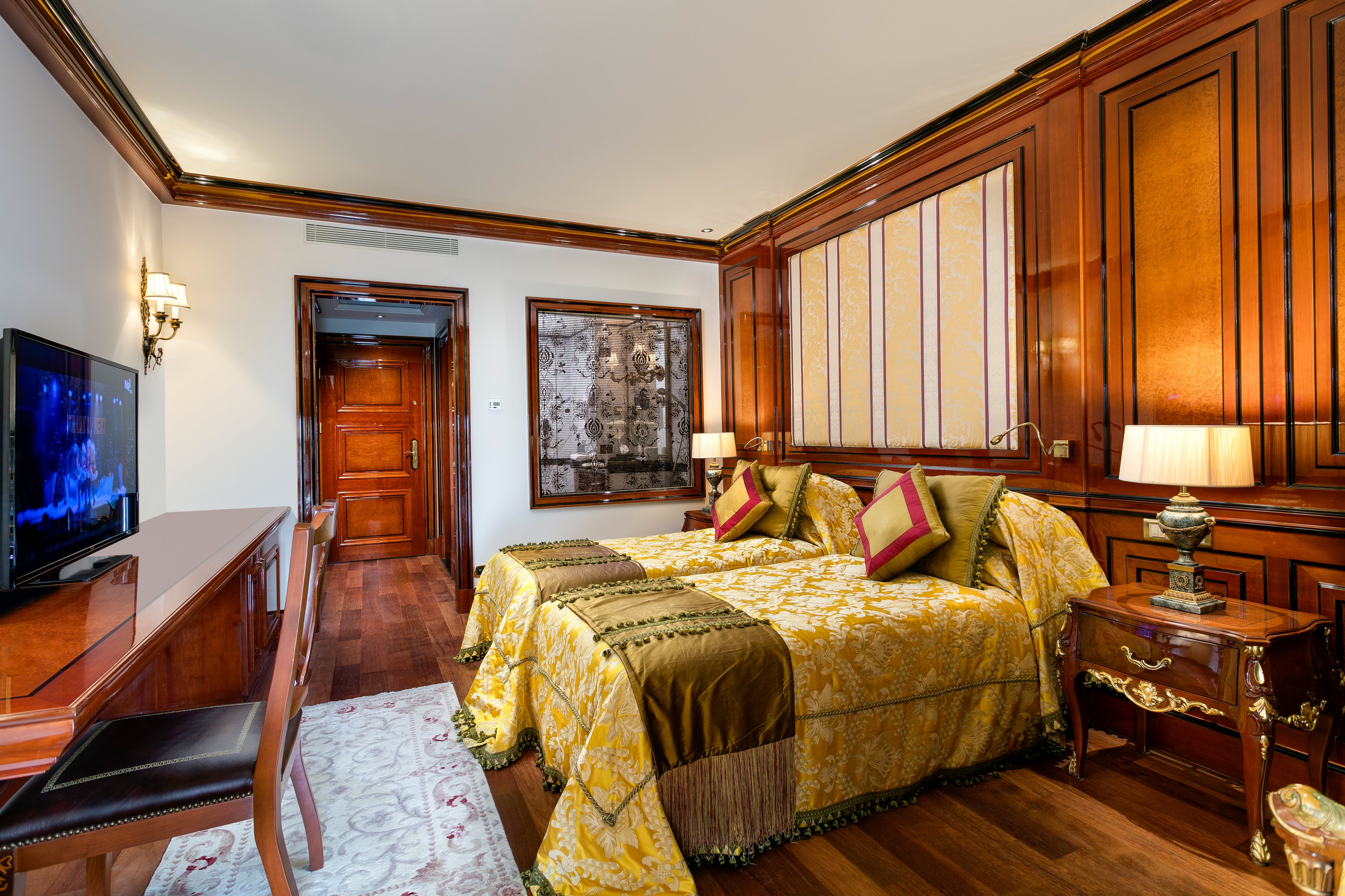 A beautifully designed hotel room featuring a double bed with golden and yellow bedding, complemented by rich wooden paneling and ornate furniture. The setting includes a desk with a chair, a television, and warm lighting from bedside lamps. The room exudes a classic and opulent style, perfect for a comfortable stay.