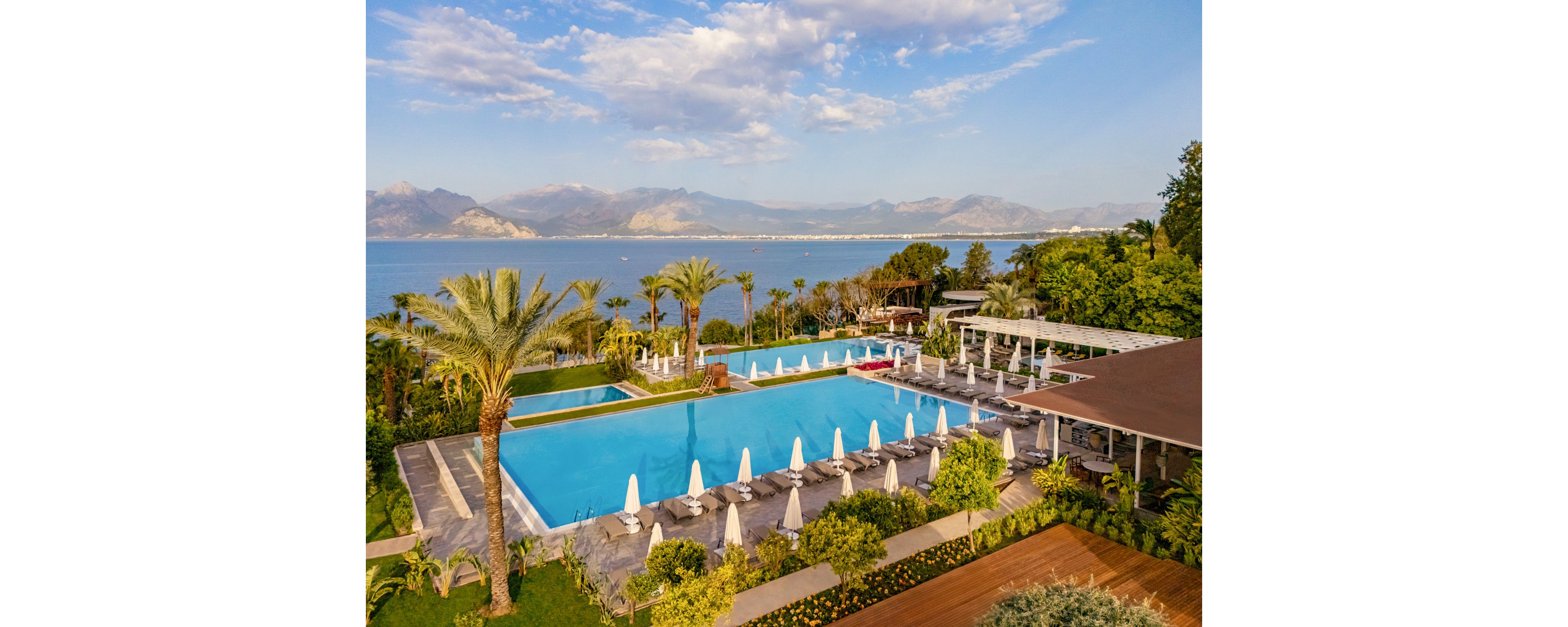 A large luxury resort features expansive infinity-style swimming pools surrounded by sun loungers and white umbrellas. Palm trees and lush landscaped gardens frame the pool area, creating a tropical atmosphere. Beyond the resort, a calm blue sea stretches toward distant mountains under a partly cloudy sky. The scene suggests a tranquil vacation destination focused on relaxation and scenic coastal views.