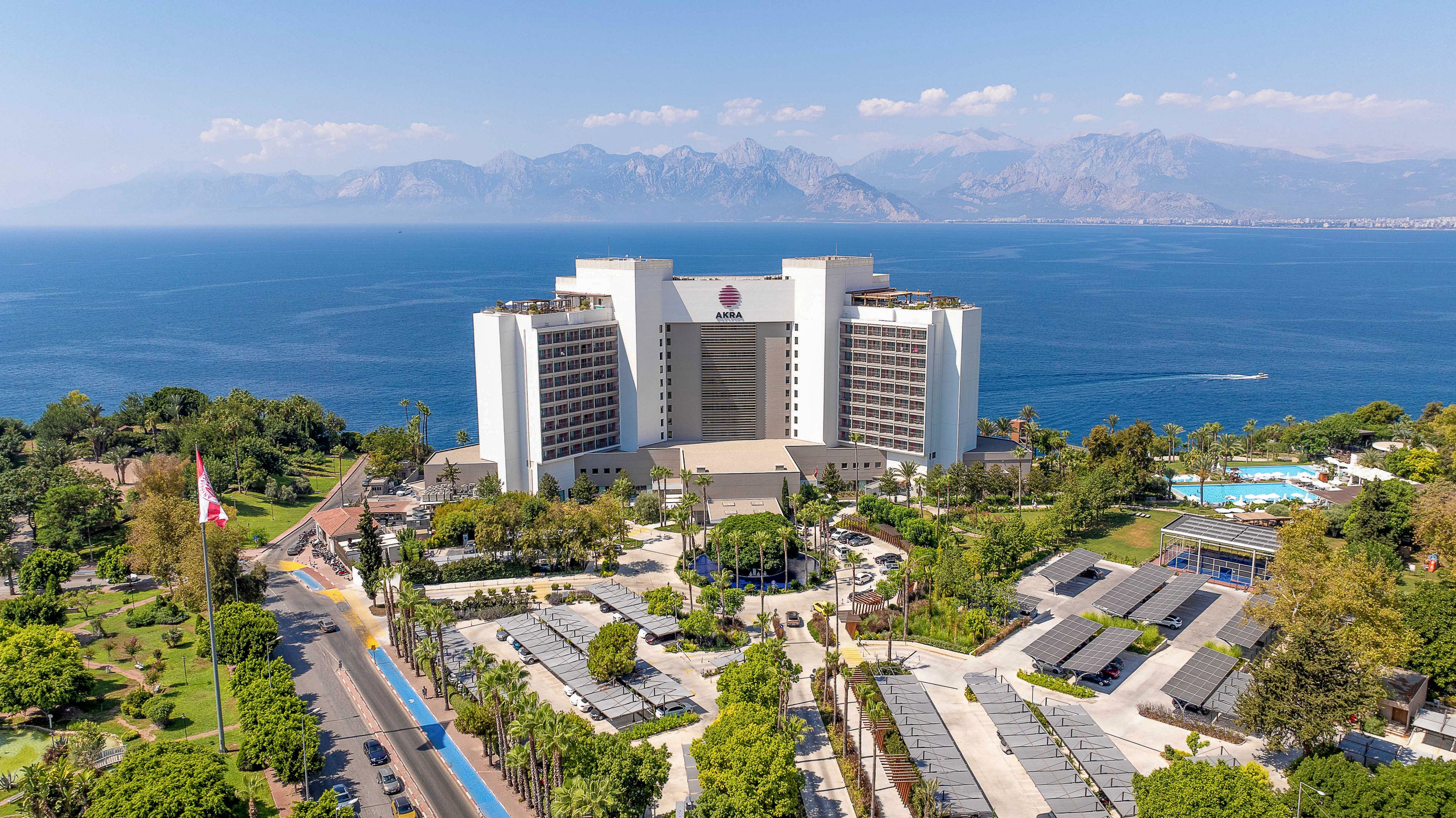 A large modern resort hotel stands prominently near the coastline, facing a calm blue sea. The property is surrounded by lush greenery, outdoor pools, and landscaped walkways. Solar panel arrays and parking areas are visible in the foreground, with a road running alongside the complex. In the distance, a dramatic mountain range rises under a clear, bright sky, emphasizing the scenic coastal setting.
