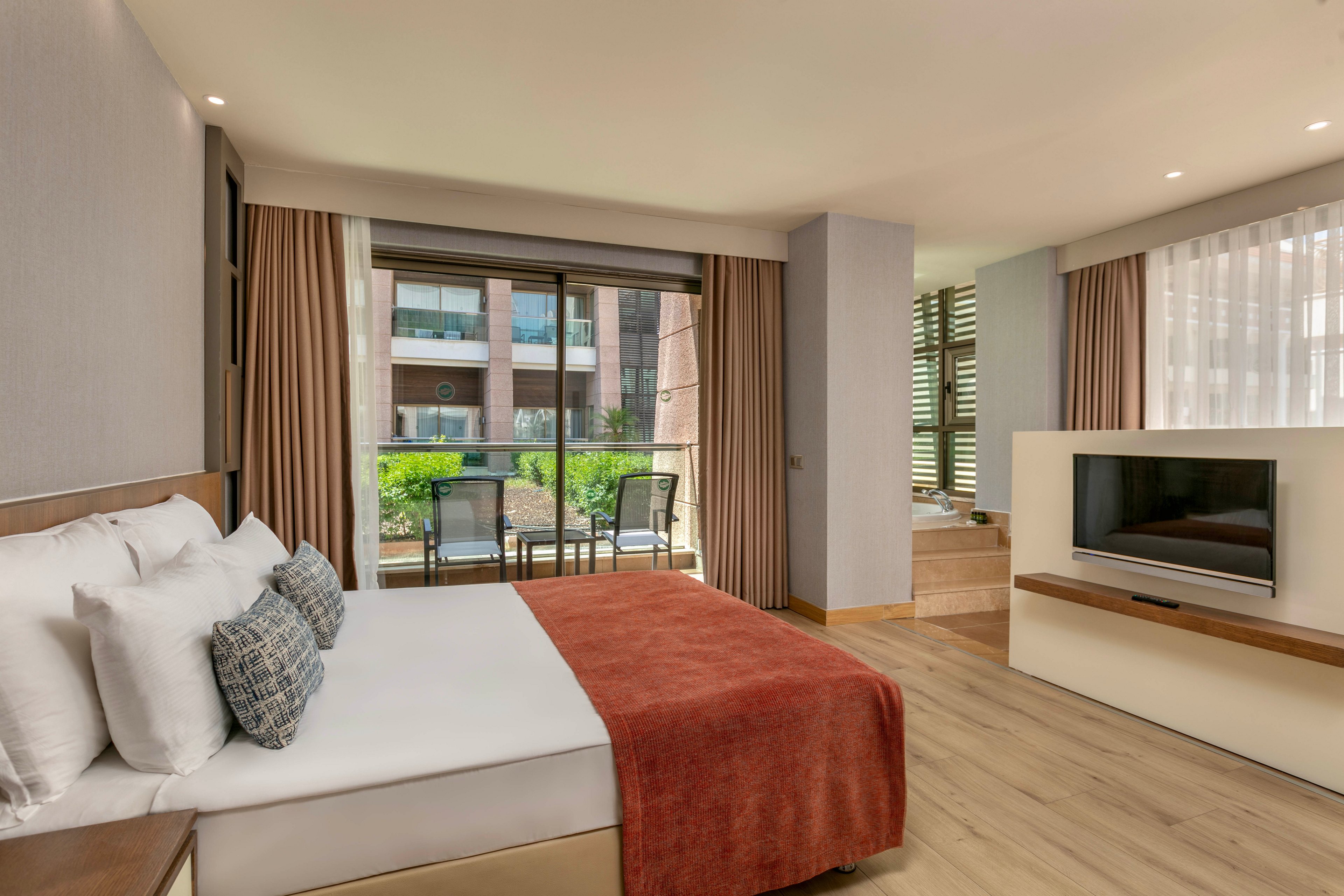 A contemporary hotel bedroom features a neatly made bed with white pillows and a rust-colored throw blanket. Floor-to-ceiling windows with beige curtains open onto a balcony overlooking a landscaped courtyard and neighboring buildings. A flat-screen television is mounted on a low partition, separating the sleeping area from a small workspace or sitting area. Soft neutral tones, wooden flooring, and natural light create a calm and inviting atmosphere.