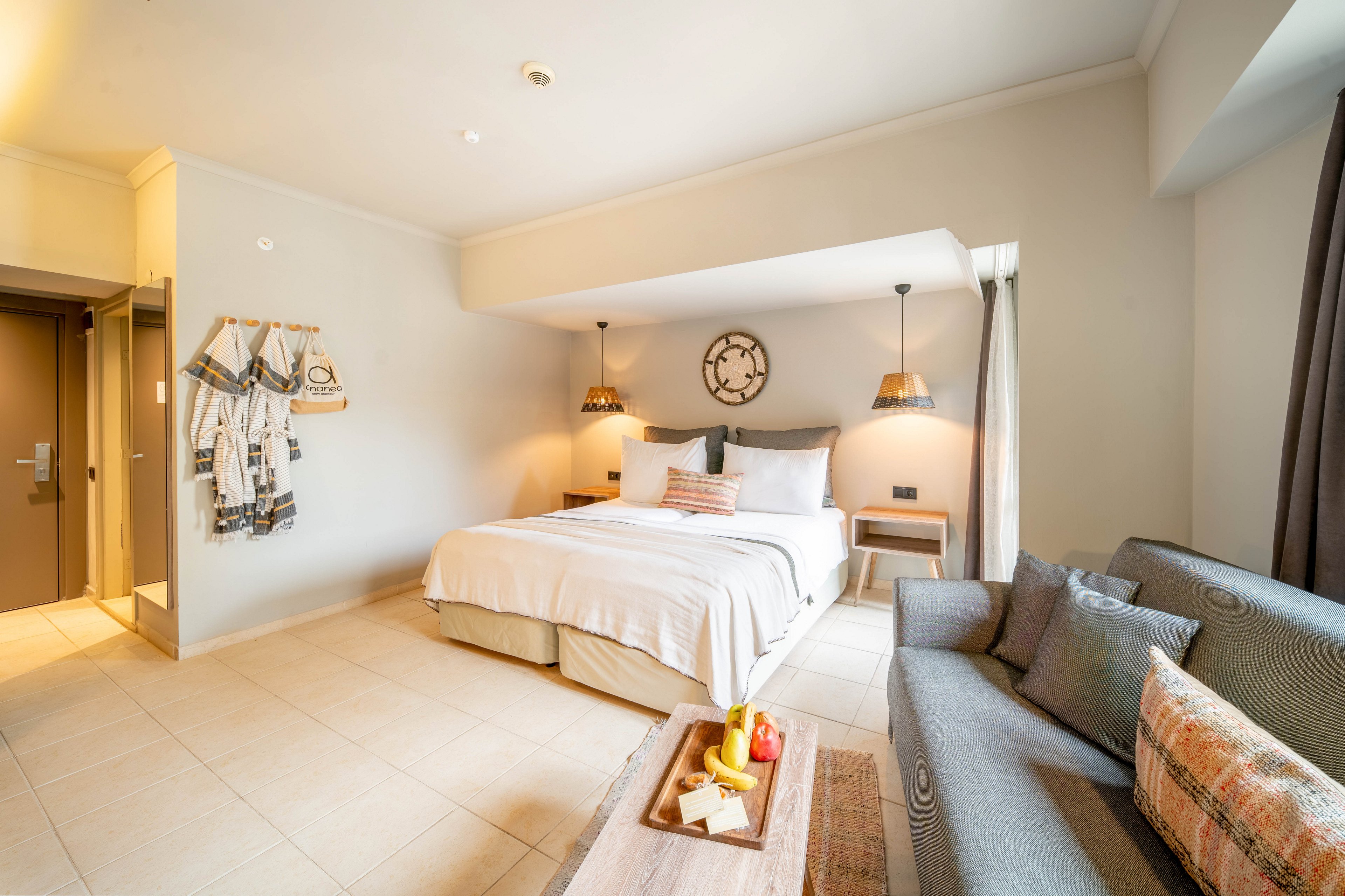 A spacious, modern hotel room features a large bed with white linens and warm accent pillows. Soft pendant lights and a decorative wall clock create a cozy atmosphere above the bed. A comfortable sofa sits opposite, with a wooden coffee table holding a tray of fruit and refreshments. Neutral tones, clean lines, and natural light from a window contribute to the inviting, contemporary interior design.