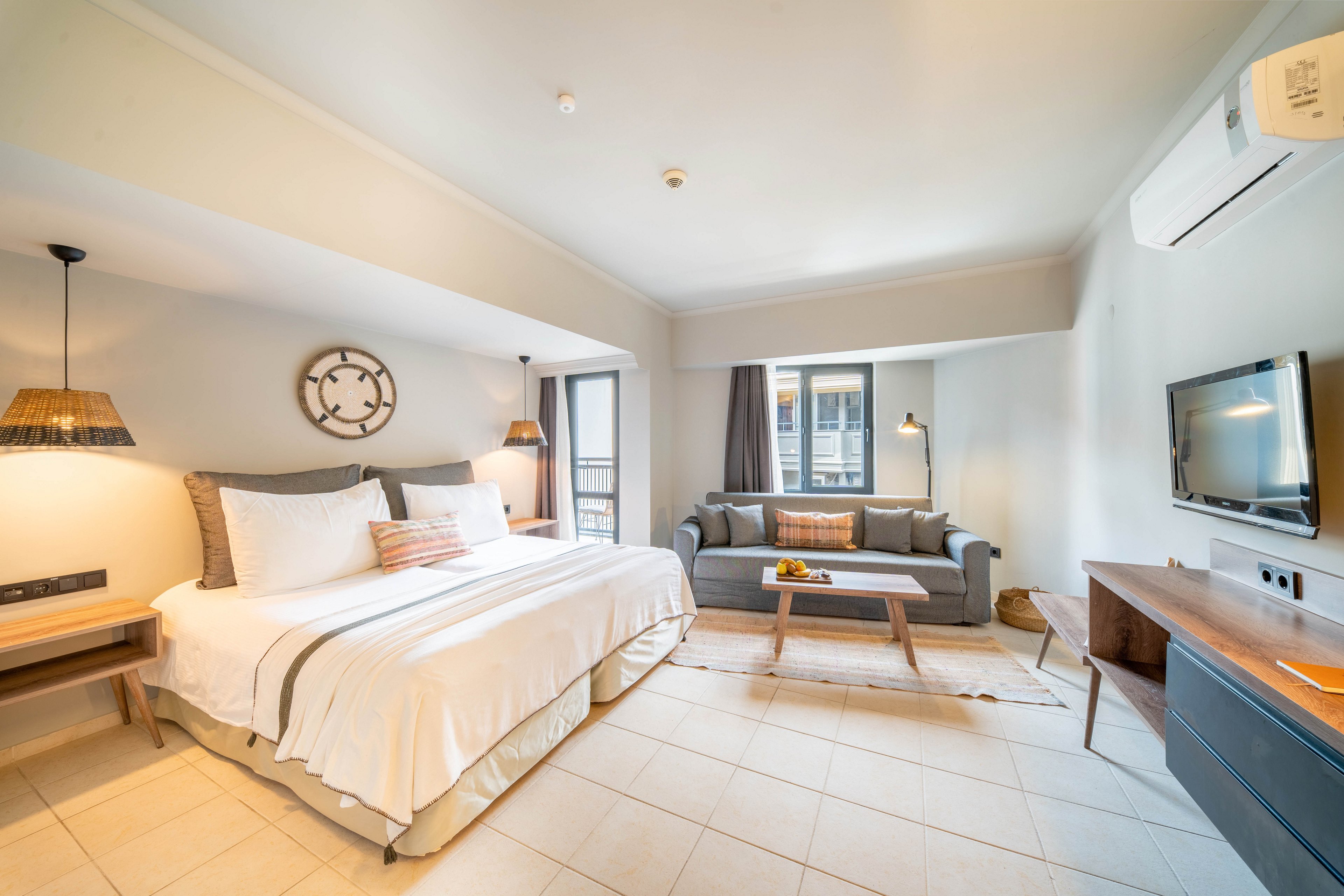 A spacious, modern hotel room features a large bed with white linens and soft accent pillows. A gray sofa and wooden coffee table create a comfortable seating area near the windows. Warm pendant lighting, neutral walls, and tiled flooring give the space a clean, contemporary feel. A wall-mounted television and air conditioning unit complete the functional, inviting interior.