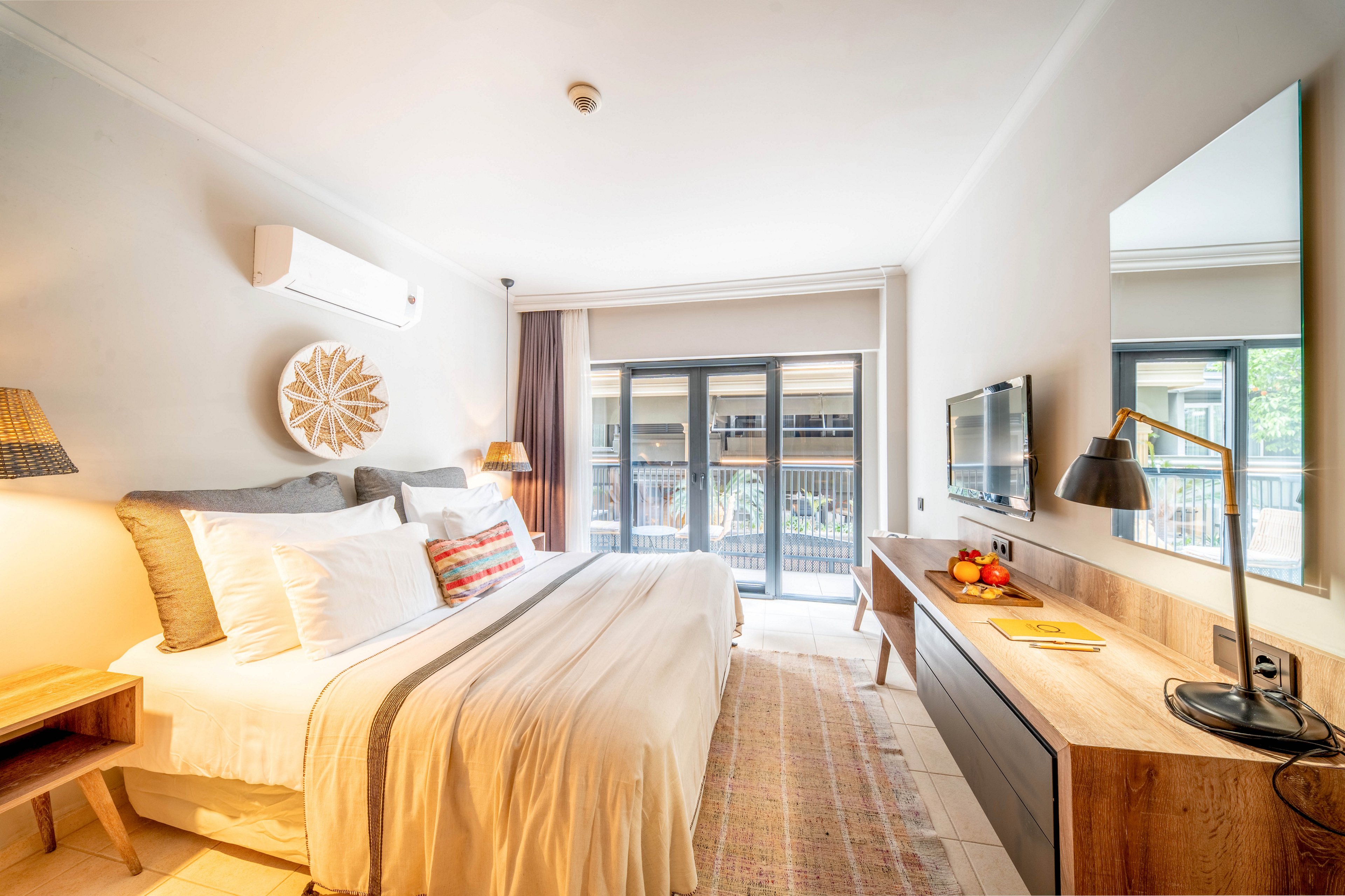 A bright modern hotel bedroom features a large neatly made bed with layered pillows and a neutral bedspread. The room includes a wooden nightstand, a long wooden desk with a lamp, and a wall-mounted television. Sliding glass doors open to a balcony with railings and greenery visible outside. Warm lighting, soft textiles, and light-colored walls create an inviting and cozy atmosphere.