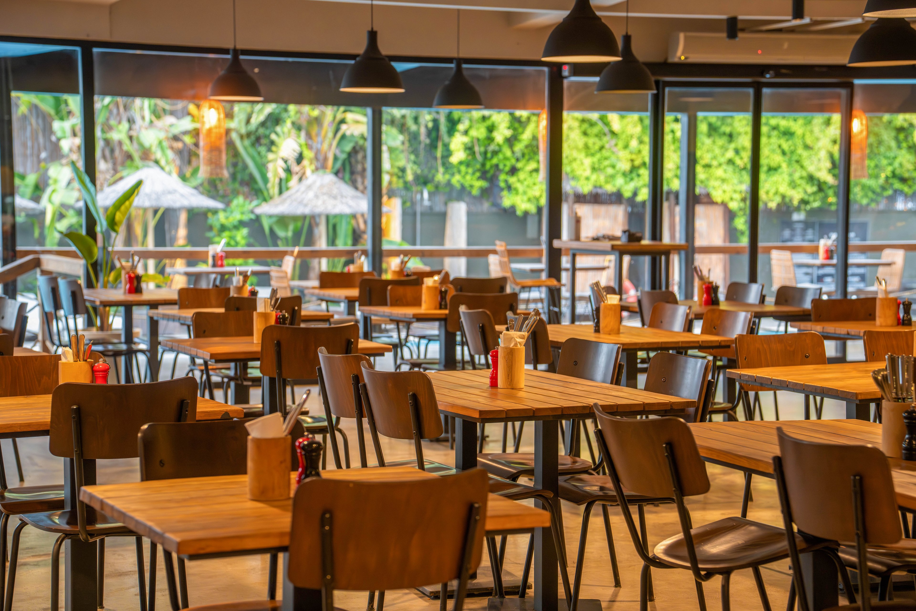 A contemporary restaurant dining area features rows of wooden tables and chairs neatly arranged. Large floor-to-ceiling windows reveal lush greenery and outdoor umbrellas on a patio. Warm pendant lights hang from the ceiling, creating a cozy and inviting atmosphere despite the absence of people. Condiment bottles and small containers are placed on each table, adding subtle color accents to the neutral interior.