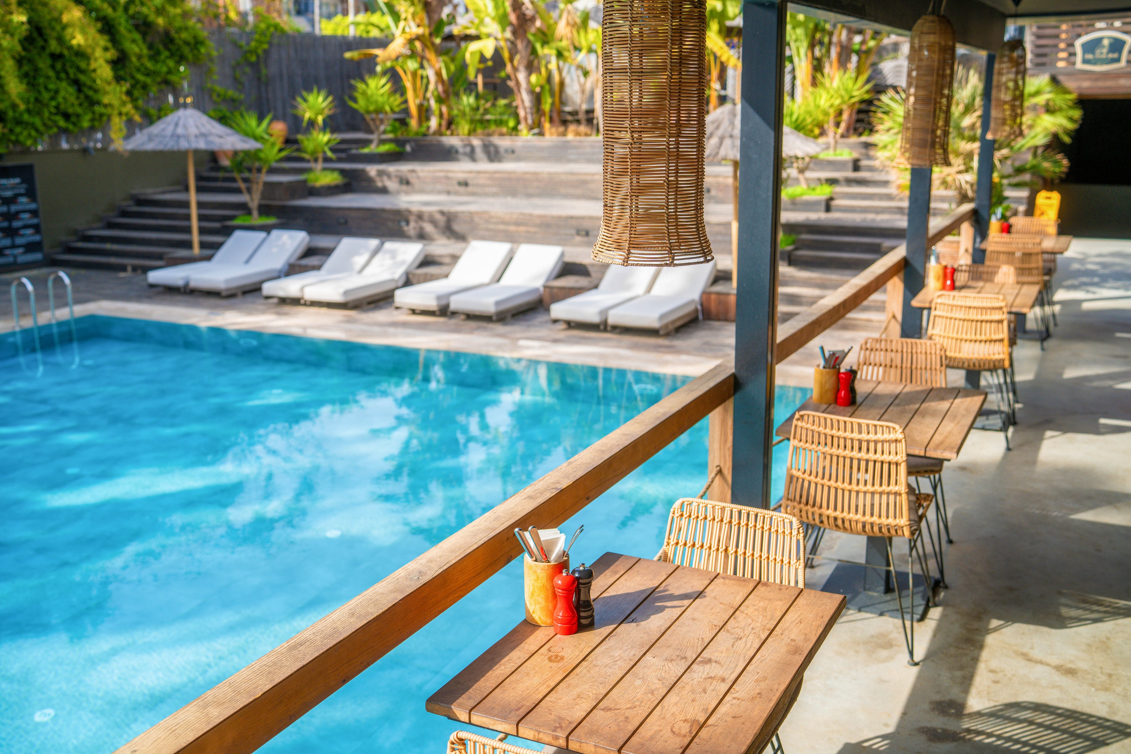An inviting poolside bar terrace overlooks a clear blue swimming pool. Wooden tables with wicker chairs line the railing, each set with drinks and utensils. In the background, several white sun loungers are arranged on stepped decking beside tropical plants. Warm sunlight enhances the relaxed resort atmosphere and highlights the natural materials and bright colors.
