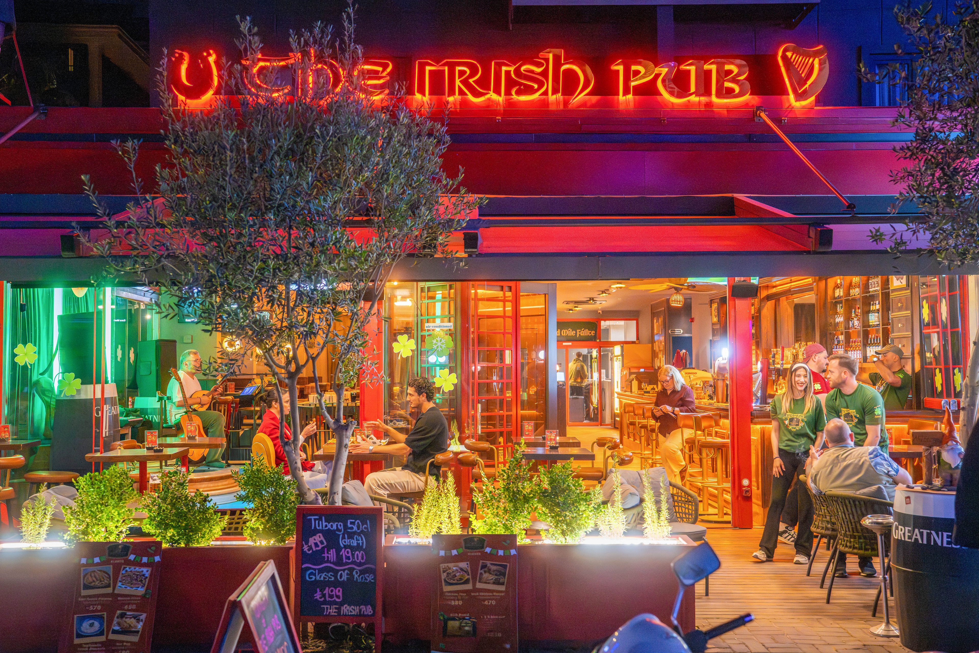 A vibrant Irish pub glows at night with bright neon signage and colorful lighting. Patrons sit at tables inside and on the terrace, enjoying drinks and conversation. The warm interior lighting contrasts with green and red exterior lights, creating a festive atmosphere. A group of people stands near the entrance, adding to the lively social scene.