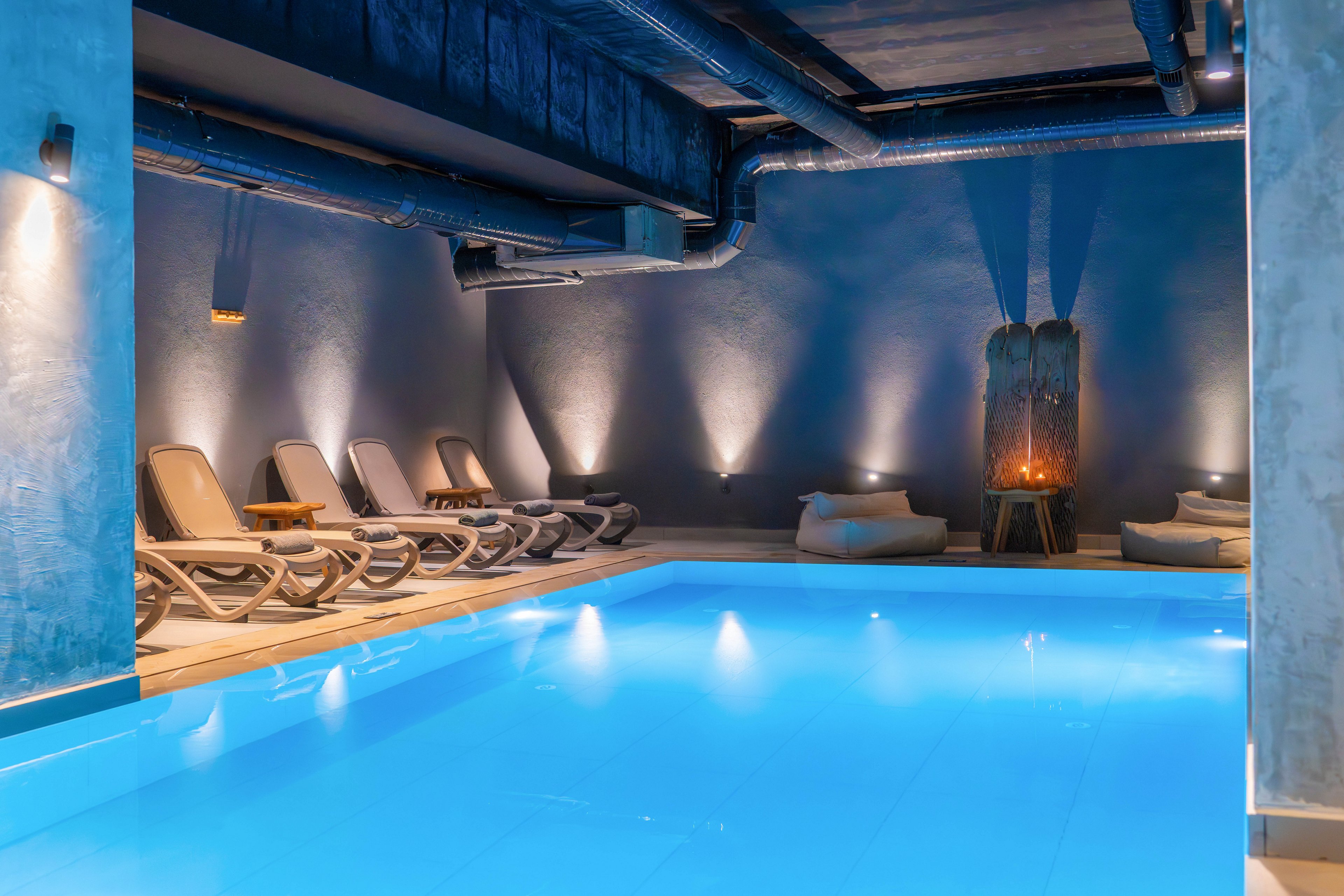 A serene indoor swimming pool glows with soft blue lighting in a contemporary spa setting. Several reclining lounge chairs line the side of the pool, inviting relaxation. Warm wall lights and a decorative fire feature create a cozy contrast against the cool-toned walls. Exposed ceiling pipes and minimalist decor give the space a modern, industrial feel.