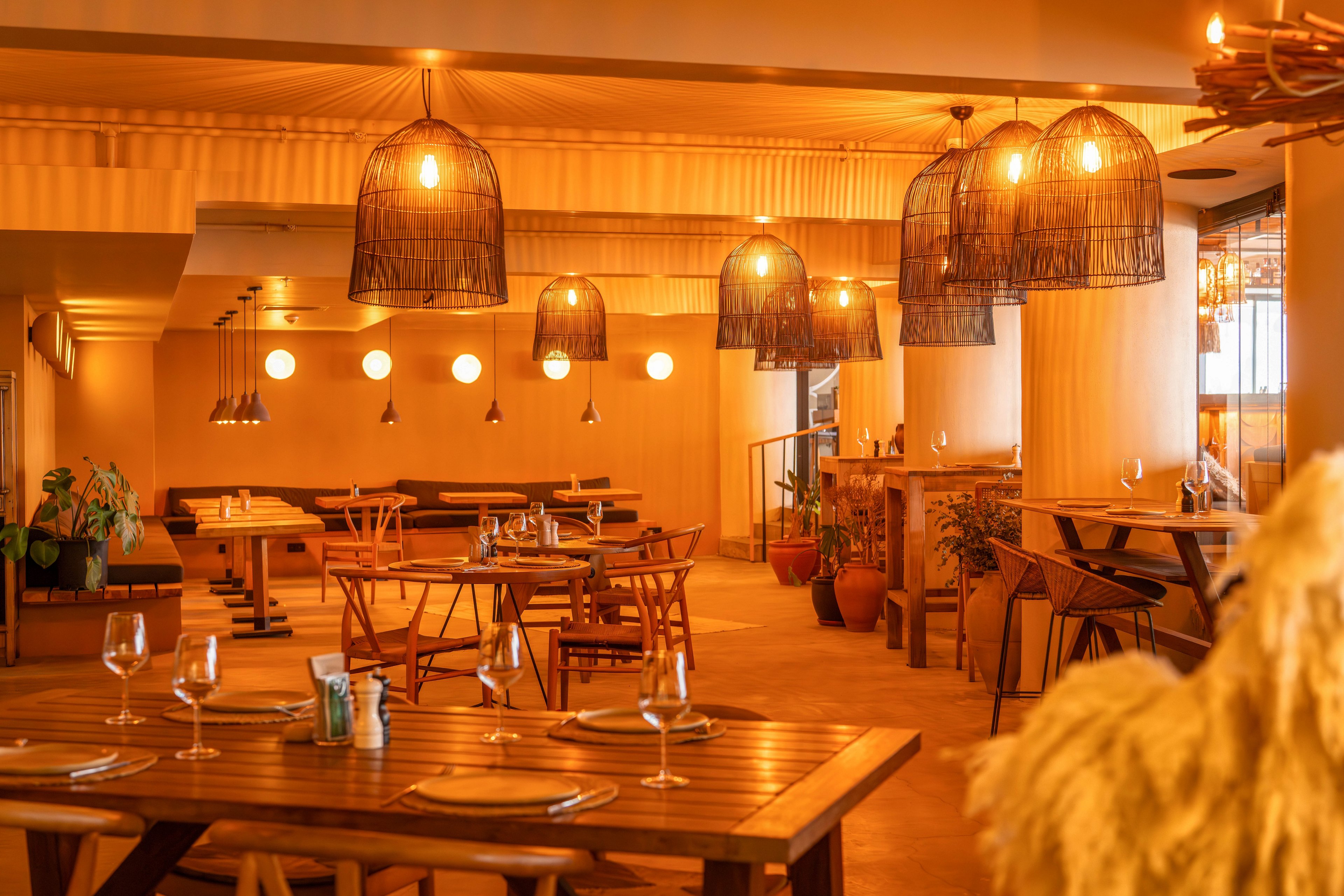 A warmly lit restaurant interior features wooden tables and chairs arranged neatly in an inviting dining area. Hanging wicker pendant lamps cast a soft amber glow across the room, highlighting the natural wood tones. Empty wine glasses and place settings are prepared on the tables, suggesting readiness for guests. Potted plants and subtle decorative elements add a relaxed, contemporary atmosphere to the space.