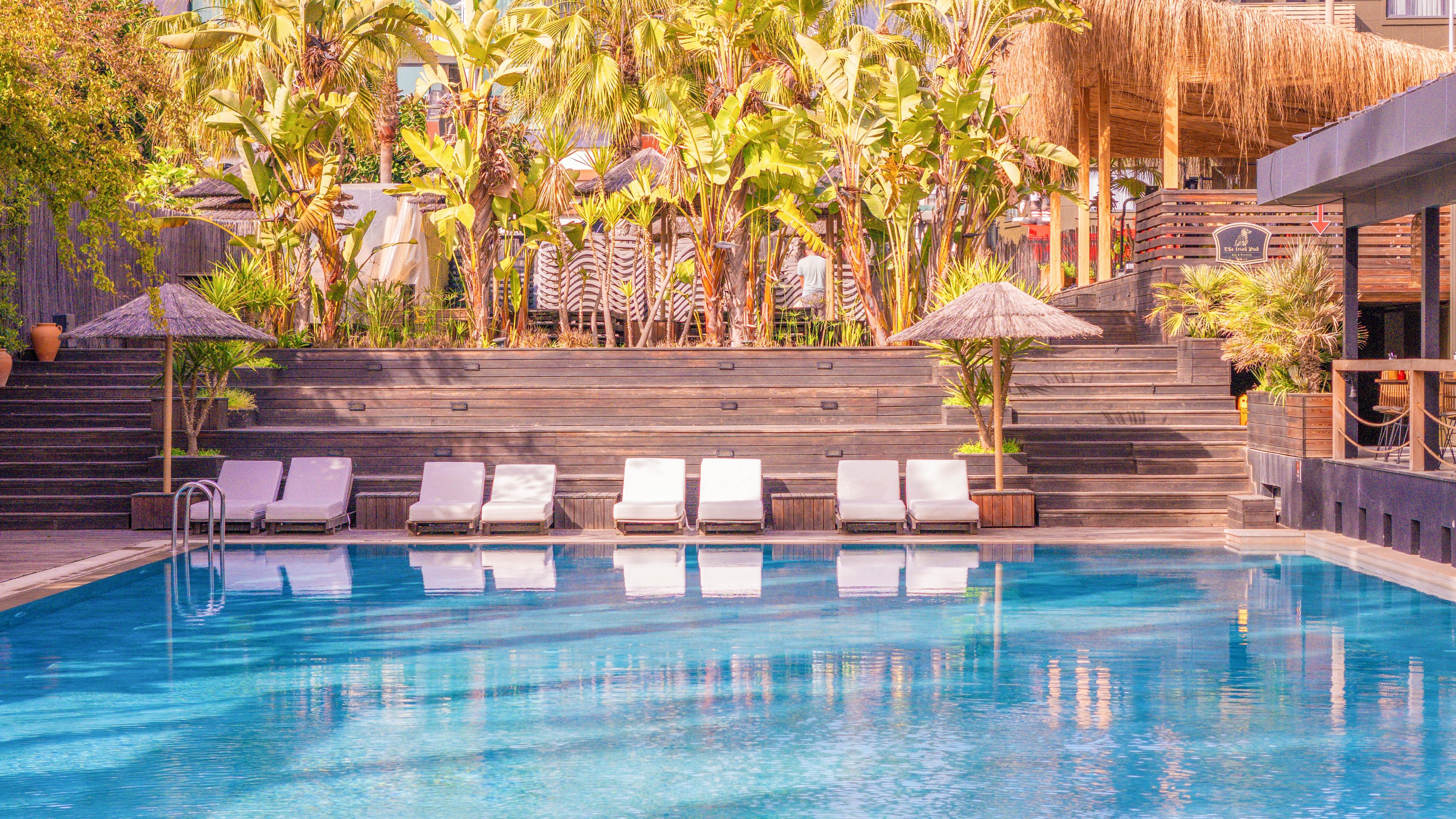 A clear blue swimming pool is set in front of a row of white sun loungers and umbrellas. The scene is framed by dark wooden decking and steps leading up to lush tropical palm trees. A thatched-roof structure and modern building elements are visible in the background, suggesting a stylish resort setting. Warm sunlight and soft reflections on the water create a relaxed vacation atmosphere.