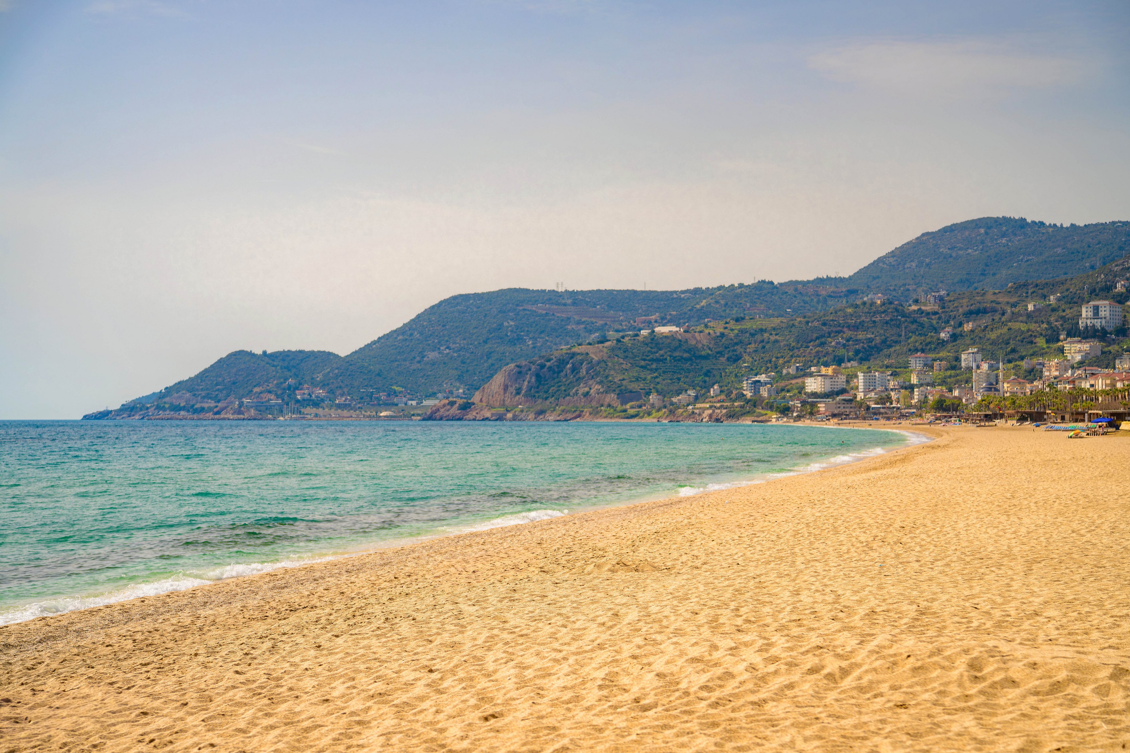 A wide sandy beach stretches along a calm turquoise sea under a hazy sky. Gentle waves roll onto the shore, leaving subtle patterns in the sand. In the distance, green hills rise above a coastal town with scattered buildings. The scene feels quiet and peaceful, suggesting a warm day by the seaside.