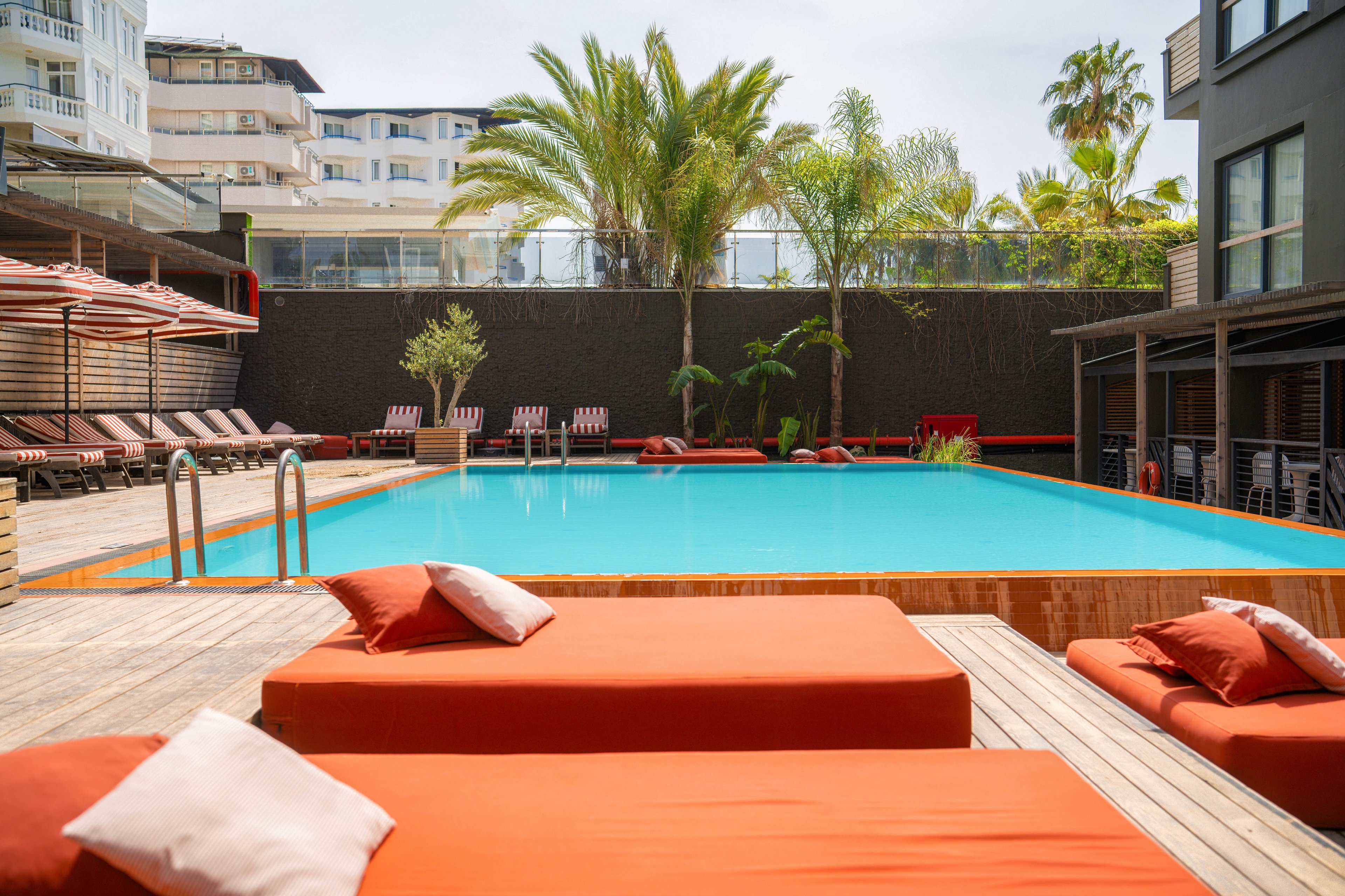 A contemporary outdoor swimming pool is surrounded by bright orange cushioned sun loungers and wooden decking. The scene is set in a modern hotel or resort courtyard with multi-story buildings in the background. Palm trees and tropical plants add greenery around the pool, while umbrellas and additional lounge chairs line the far side. The clear blue water and soft cushions create a relaxed, vacation atmosphere under a bright sky.