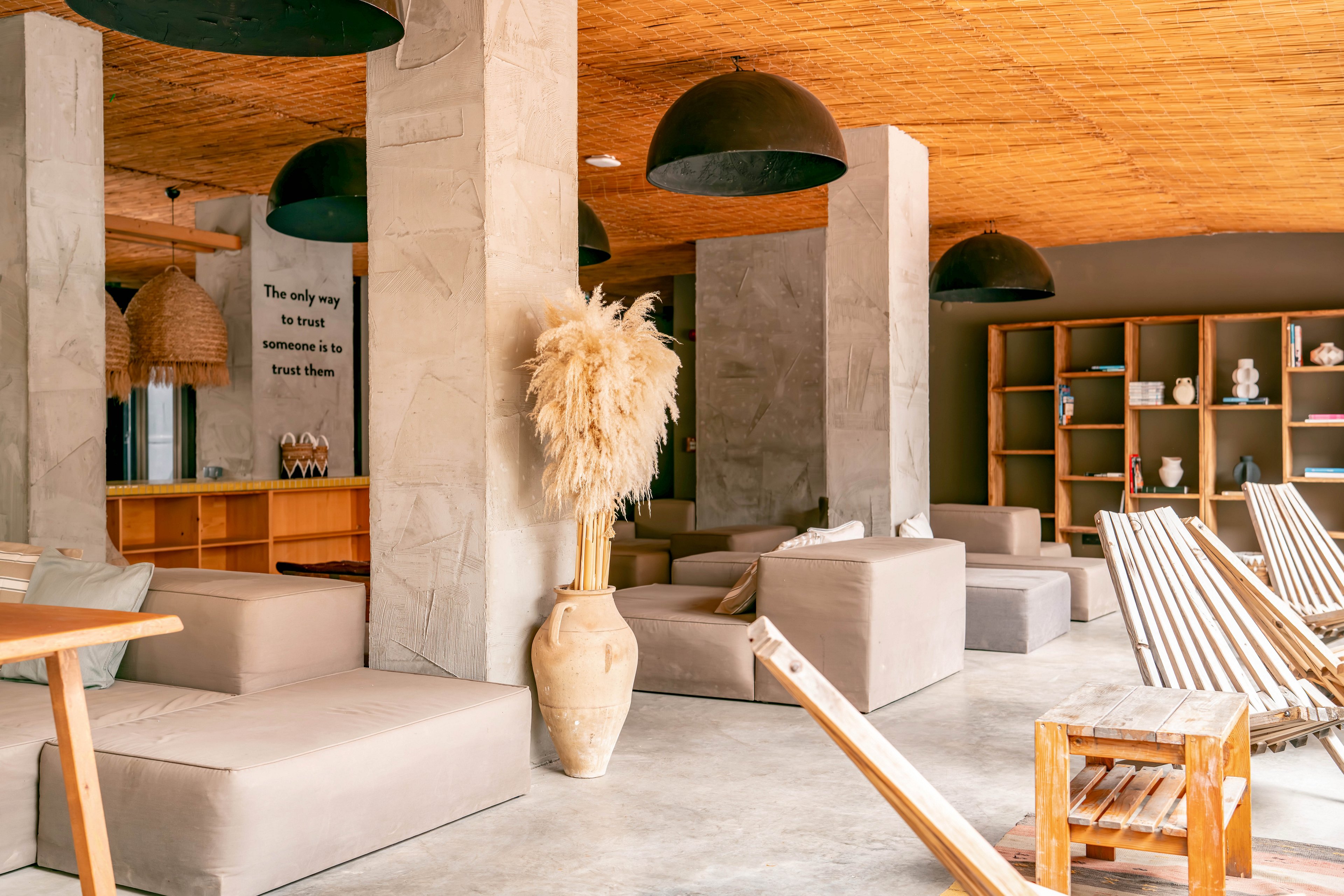 A contemporary lounge area features low beige sofas and wooden deck chairs arranged around concrete pillars. The space has a warm wooden ceiling with large black pendant lights hanging above. A tall vase filled with dried pampas grass stands prominently in the foreground. In the background, open wooden shelves display decorative ceramics and a bar counter adds to the relaxed, social atmosphere.