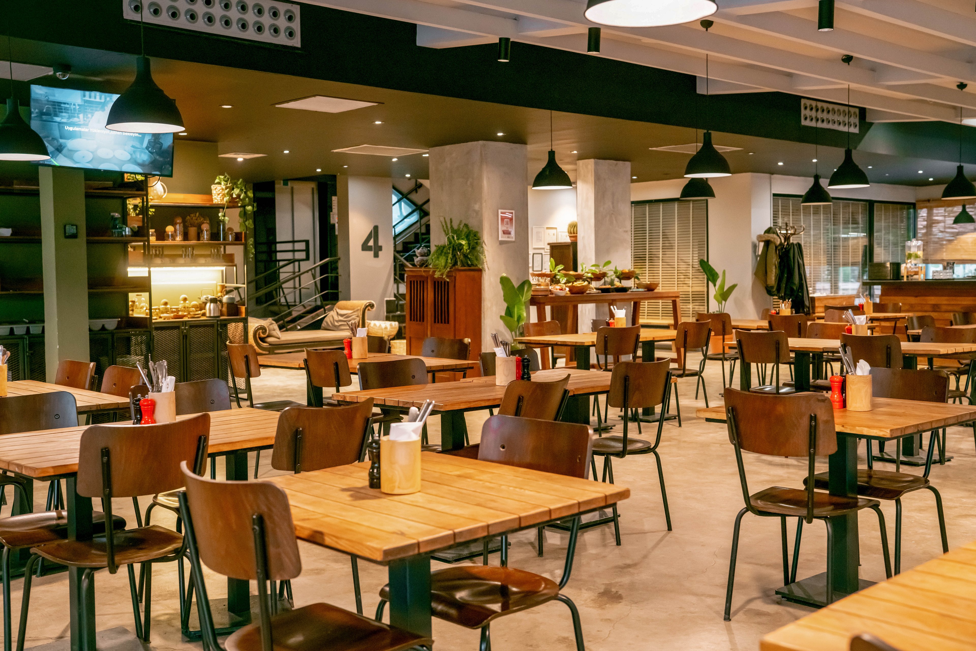 A spacious modern cafeteria features rows of wooden tables and chairs arranged neatly in an open-plan dining area. Warm pendant lights hang from the ceiling, illuminating the polished concrete floor and contemporary decor. A buffet or serving counter is visible in the background, along with shelves and a staircase leading to another level. The atmosphere feels clean, organized, and ready to welcome guests.