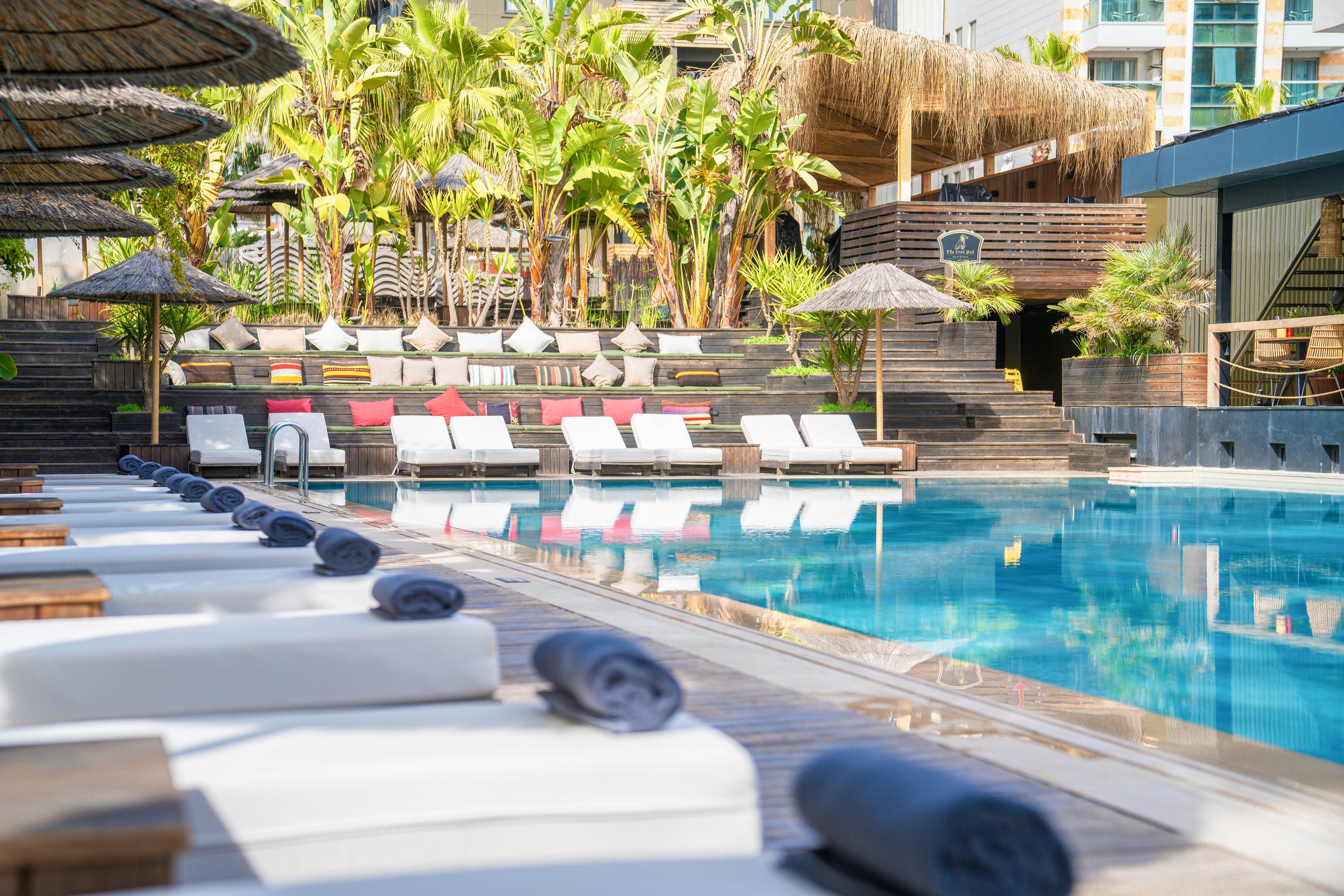 A luxurious outdoor swimming pool is surrounded by white cushioned sun loungers, each topped with neatly rolled dark towels. The scene features a wooden deck and a row of colorful cushions on seating in the background. Lush tropical plants and palm trees frame the pool area, creating a resort-like atmosphere. Modern buildings and cabana-style structures add an upscale, urban resort feel to the setting.