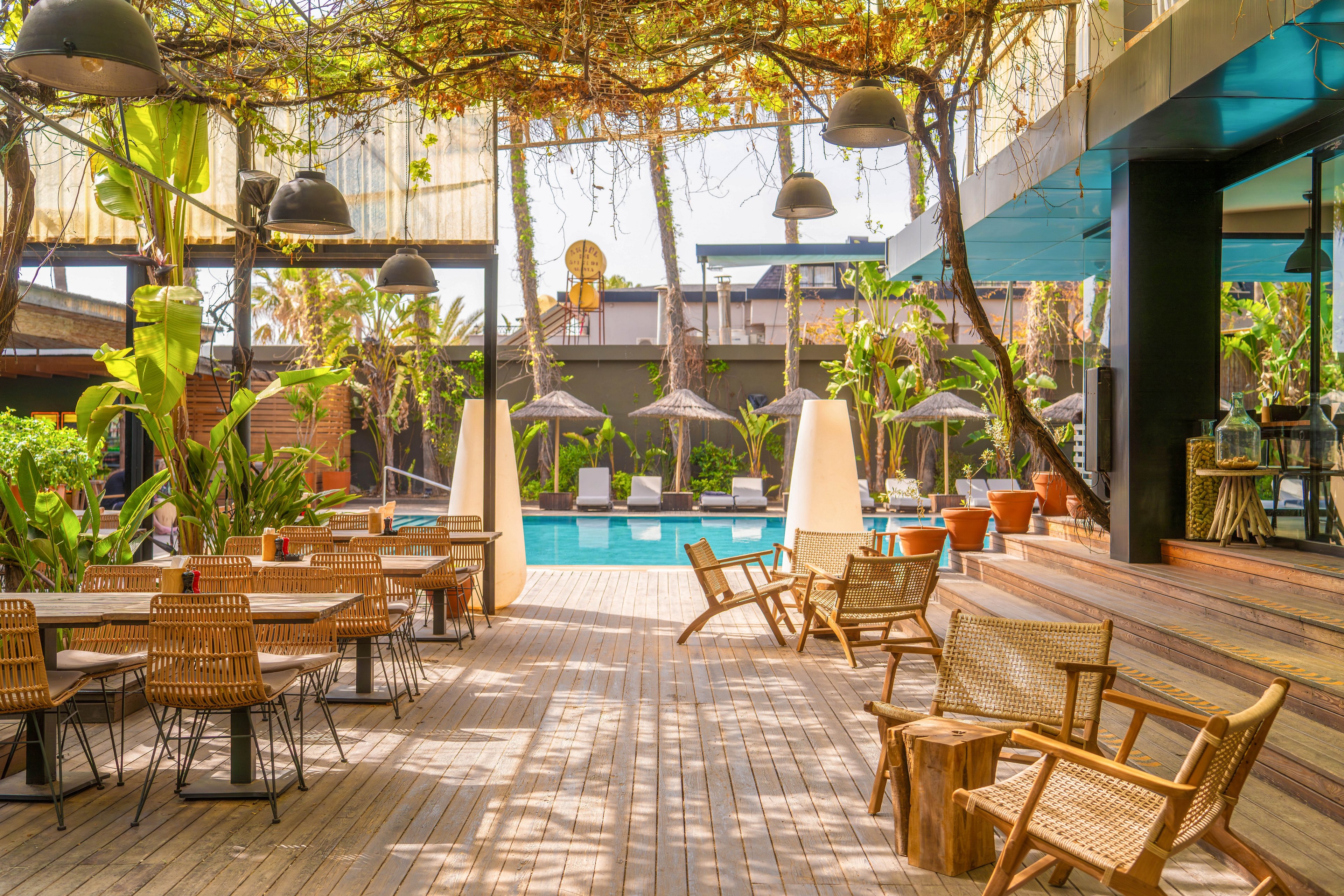 An inviting outdoor lounge area features wooden tables and woven chairs arranged on a spacious deck. In the background, a bright blue swimming pool is framed by sun loungers and large umbrellas. Lush tropical plants and hanging greenery create a shaded, resort-like atmosphere. Warm natural light and earthy tones give the scene a relaxed, vacation mood.