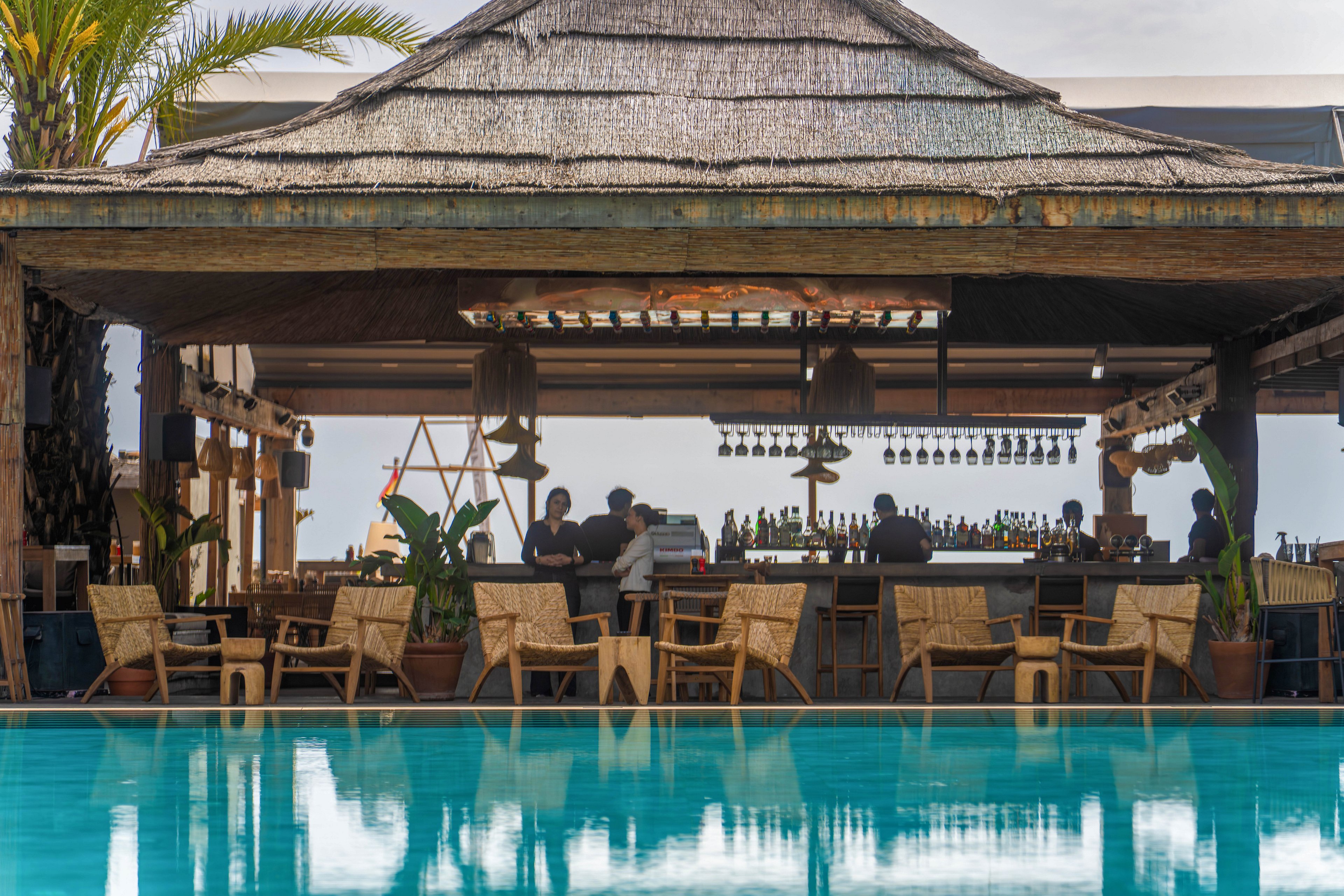 An open-air bar with a thatched roof sits beside a clear blue swimming pool. Guests and bartenders gather around the counter, with bottles and hanging glassware visible in the background. Wicker chairs and wooden tables line the poolside, creating a relaxed lounge atmosphere. Palm trees and warm lighting contribute to a tropical, resort-like setting.