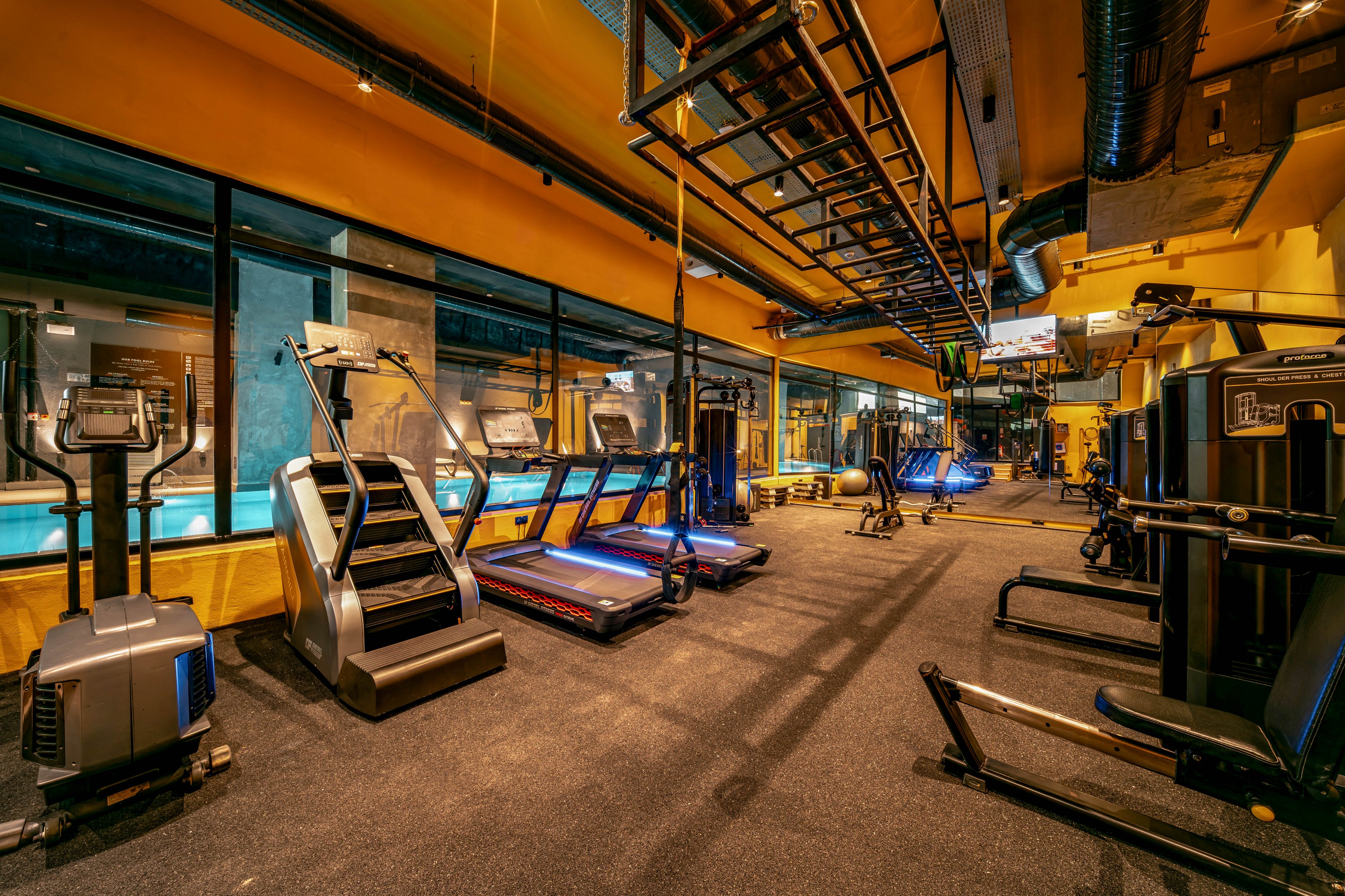 A modern fitness center features rows of treadmills and elliptical machines in a brightly lit room. The gym has a warm yellow and orange color scheme with dark flooring and ceiling fixtures. Large windows on one side reveal another area of the facility, possibly a pool or additional workout space. The equipment is powered on, but no people are present, giving the space a clean and organized appearance.