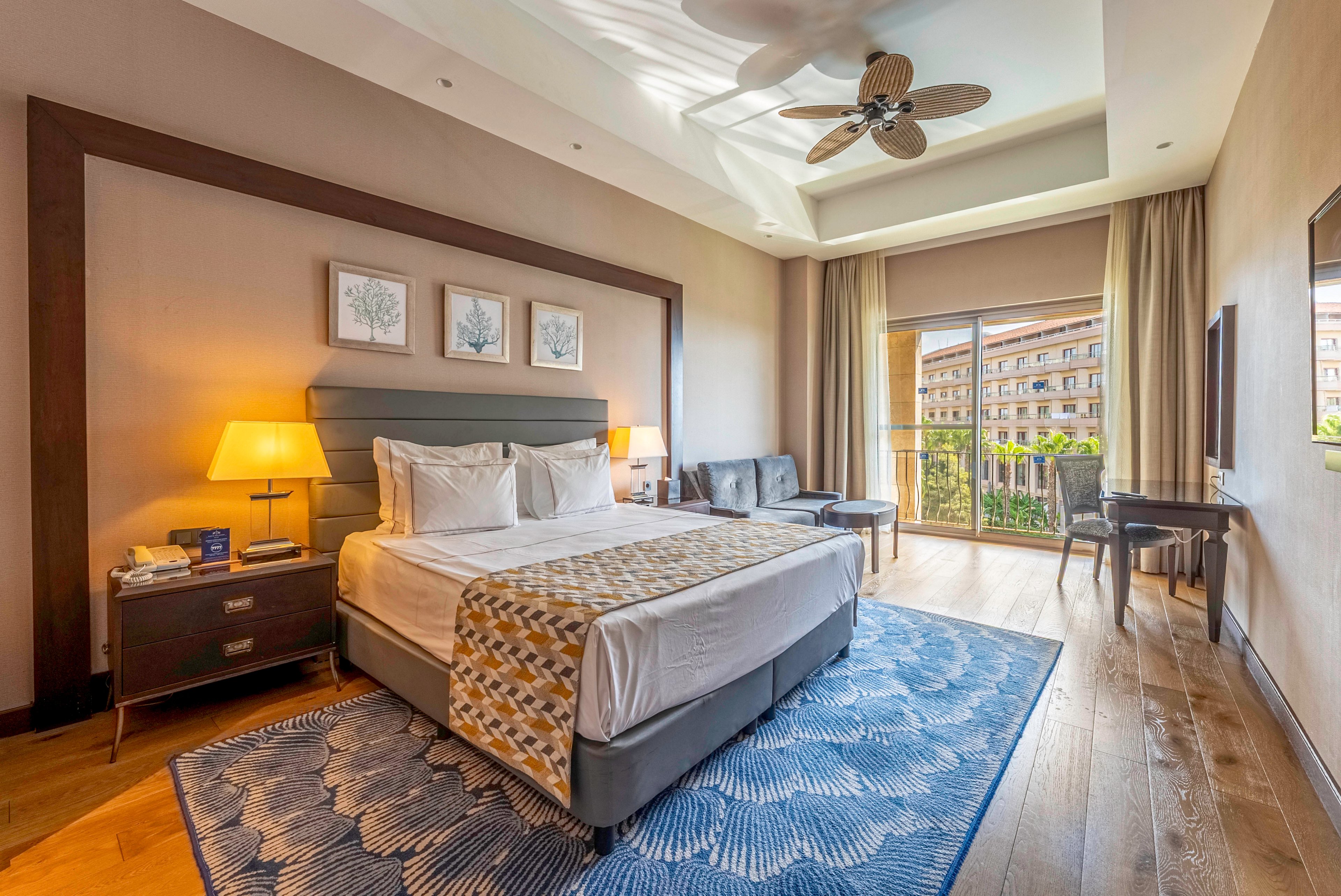 A spacious, modern hotel bedroom features a large double bed with white linens and a patterned runner. Warm lighting from a bedside lamp and natural light from floor-to-ceiling balcony doors create an inviting atmosphere. The room includes a blue area rug, wooden floors, a small sofa, and a desk with a chair. Framed artwork above the bed and a ceiling fan add decorative and functional details, while the balcony overlooks another building.