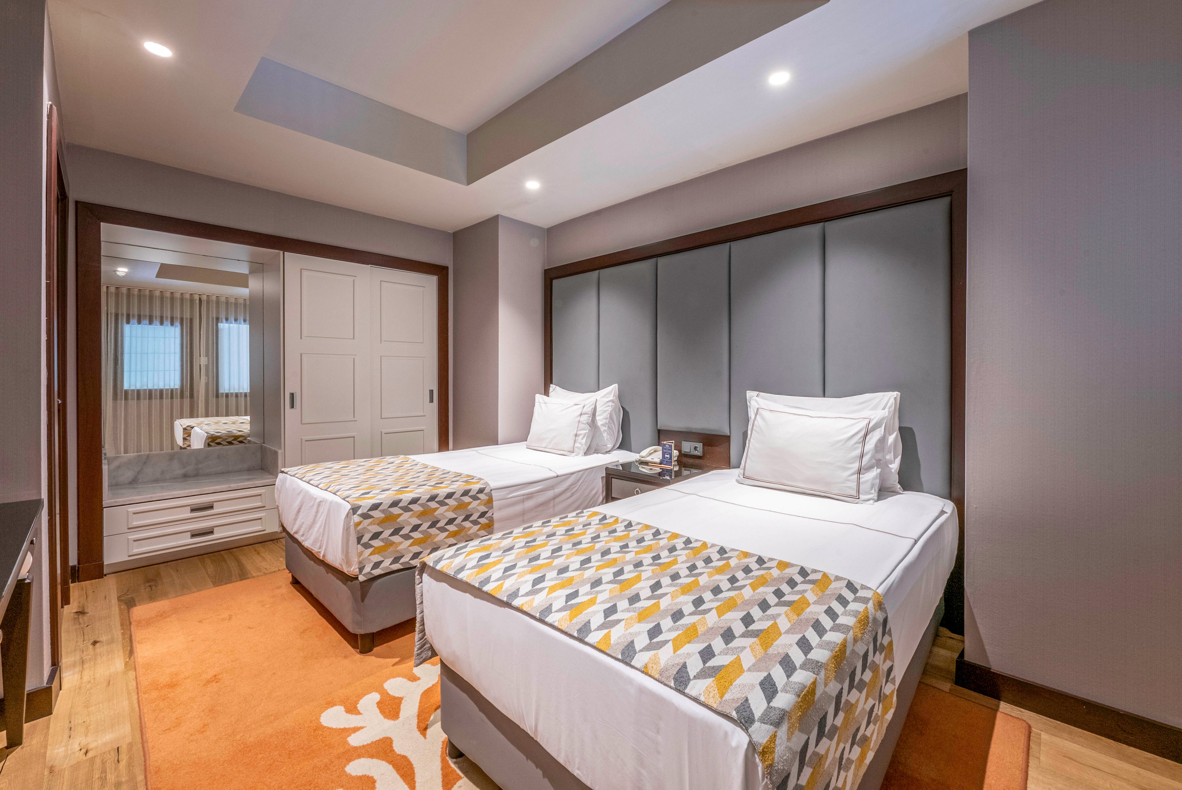 Contemporary bedroom interior featuring two neatly made twin beds with patterned bed runners. The room includes a soft orange rug, wooden flooring, and a large upholstered headboard wall. Recessed ceiling lights provide warm illumination, while a mirrored wardrobe and white double doors enhance the sense of space. Neutral tones and clean lines create a calm, inviting atmosphere suitable for guests.
