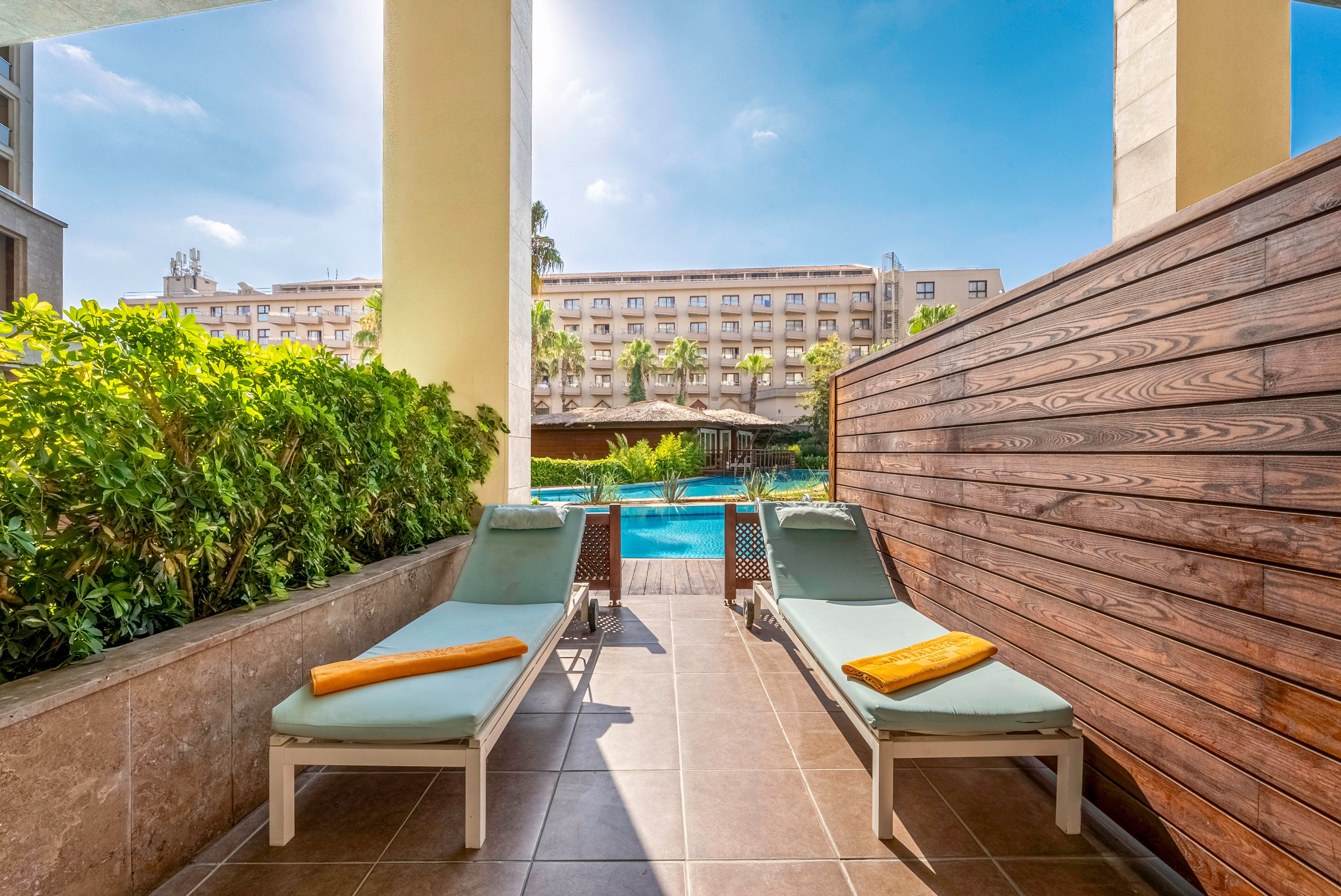 Two cushioned sun loungers with orange towels sit on a tiled private terrace overlooking a bright blue swimming pool. A wooden privacy wall and green hedges frame the space, creating a cozy outdoor nook. In the background, a large multi-story hotel building rises behind palm trees under a clear blue sky. The scene suggests a relaxing vacation setting with easy access to the pool area.