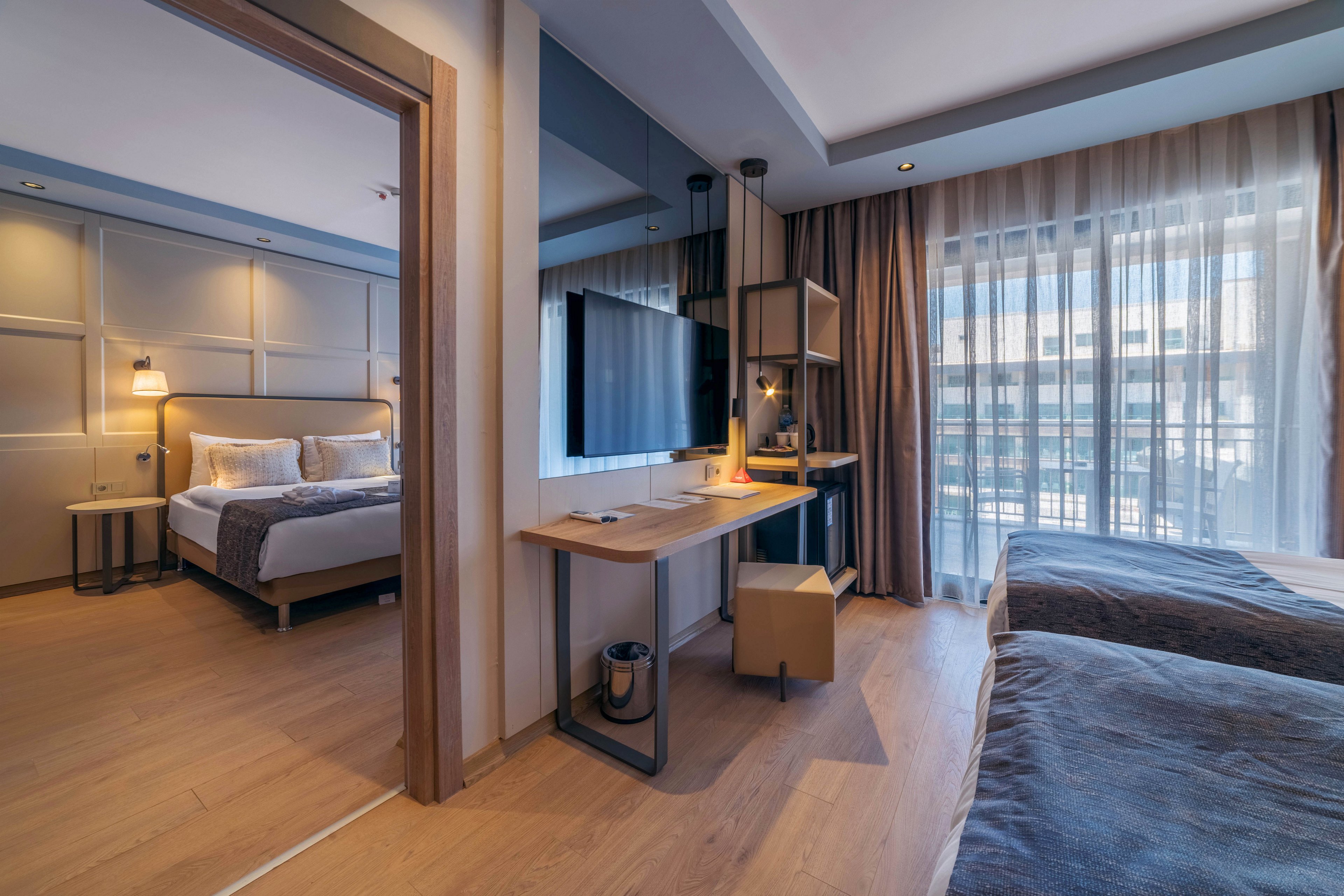 Contemporary hotel suite interior featuring a cozy bedroom with a double bed and bedside lamps. In the foreground, a small workspace with a desk, chair, and wall-mounted television separates the sleeping areas. Large floor-to-ceiling windows with sheer curtains allow natural light to fill the room and reveal an urban exterior view. Warm wood flooring and neutral-toned textiles create a calm, inviting atmosphere suitable for business or leisure travel.