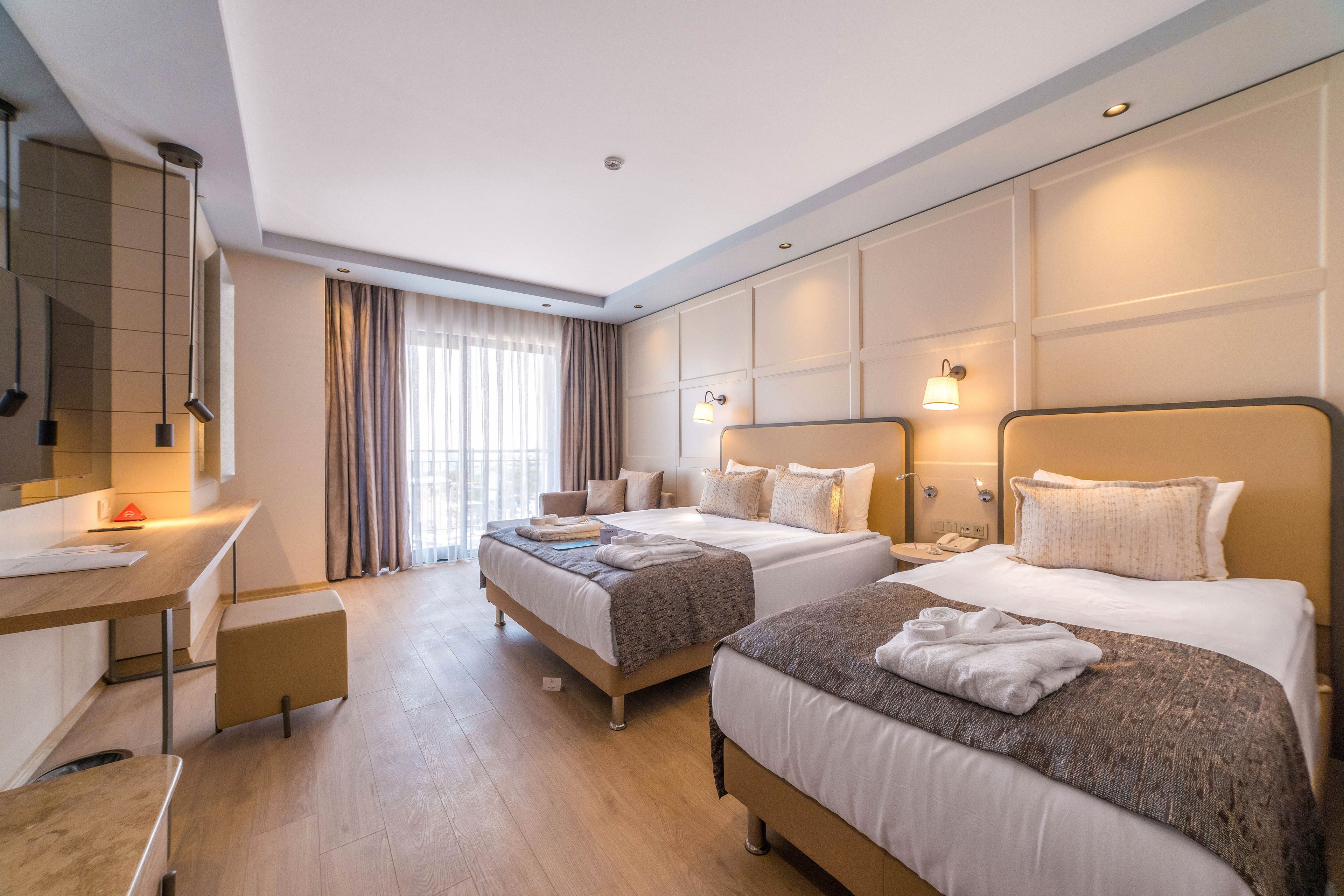 A contemporary hotel guest room features two neatly made double beds with neutral bedding and textured throws. Soft wall-mounted lights and large windows with sheer curtains create a bright, inviting atmosphere. A desk area, chair, and side tables provide functional space for work or relaxation. Warm beige tones, wood flooring, and minimalist decor contribute to a clean, comfortable, and modern interior.