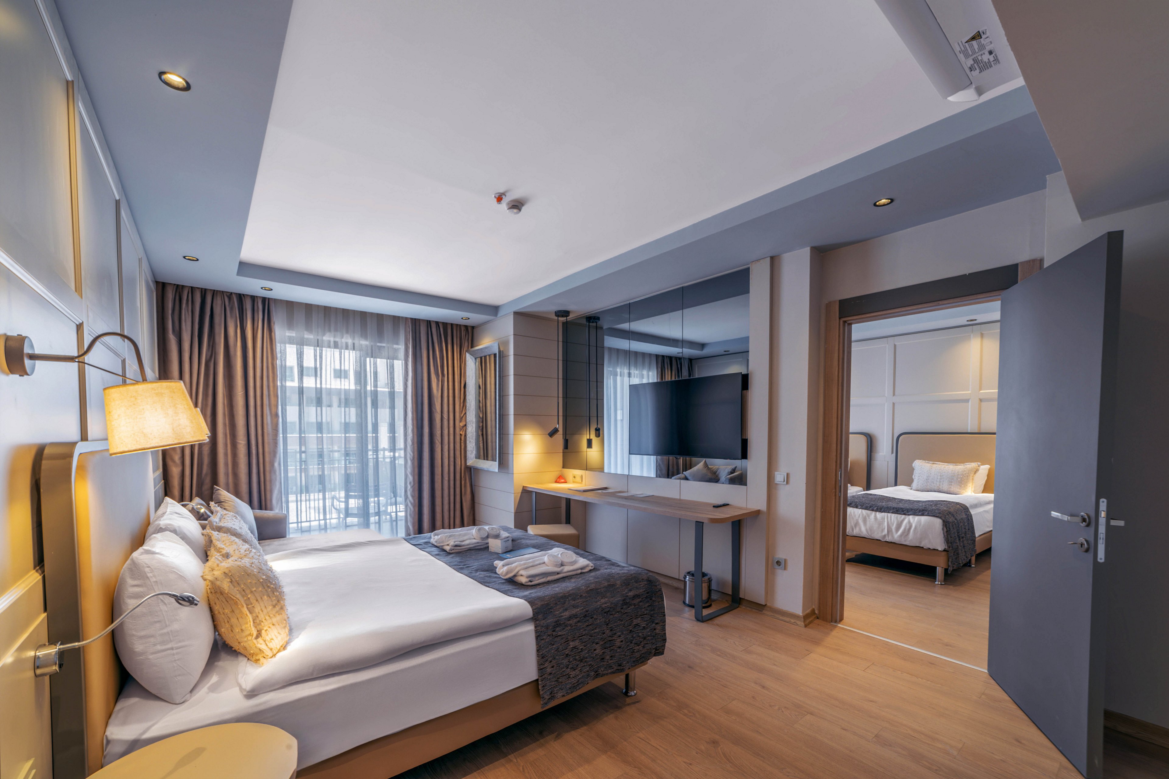 Contemporary hotel room features a neatly made double bed with plush pillows and folded towels at the foot. A large flat-screen TV is mounted on the wall above a minimalist desk, facing the main sleeping area. Floor-to-ceiling windows with sheer curtains allow natural light to fill the space, highlighting the warm wood flooring and soft neutral tones. An open door reveals an adjoining bedroom with a similar bed and lighting, suggesting a spacious suite layout.