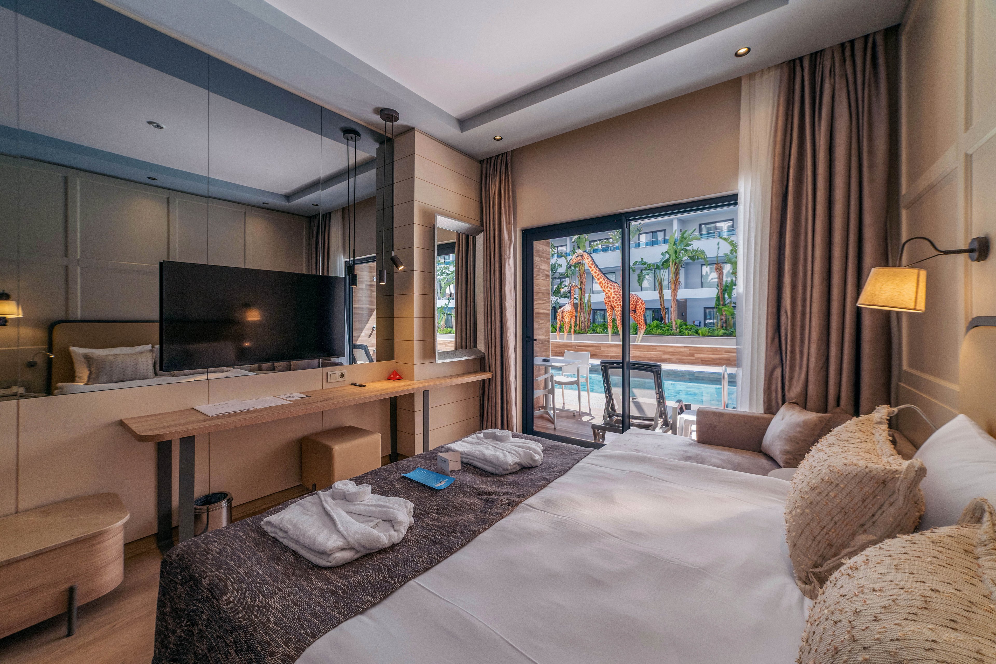 Contemporary hotel room interior with a large bed, textured pillows, and neatly folded white bathrobes on the duvet. A flat-screen TV is mounted above a long wooden desk with a chair and small items placed on top. Floor-to-ceiling curtains frame sliding glass doors that open to a balcony with seating and a view of the swimming pool and resort buildings. Warm lighting and neutral tones create a cozy, inviting atmosphere suitable for vacation or business travel.