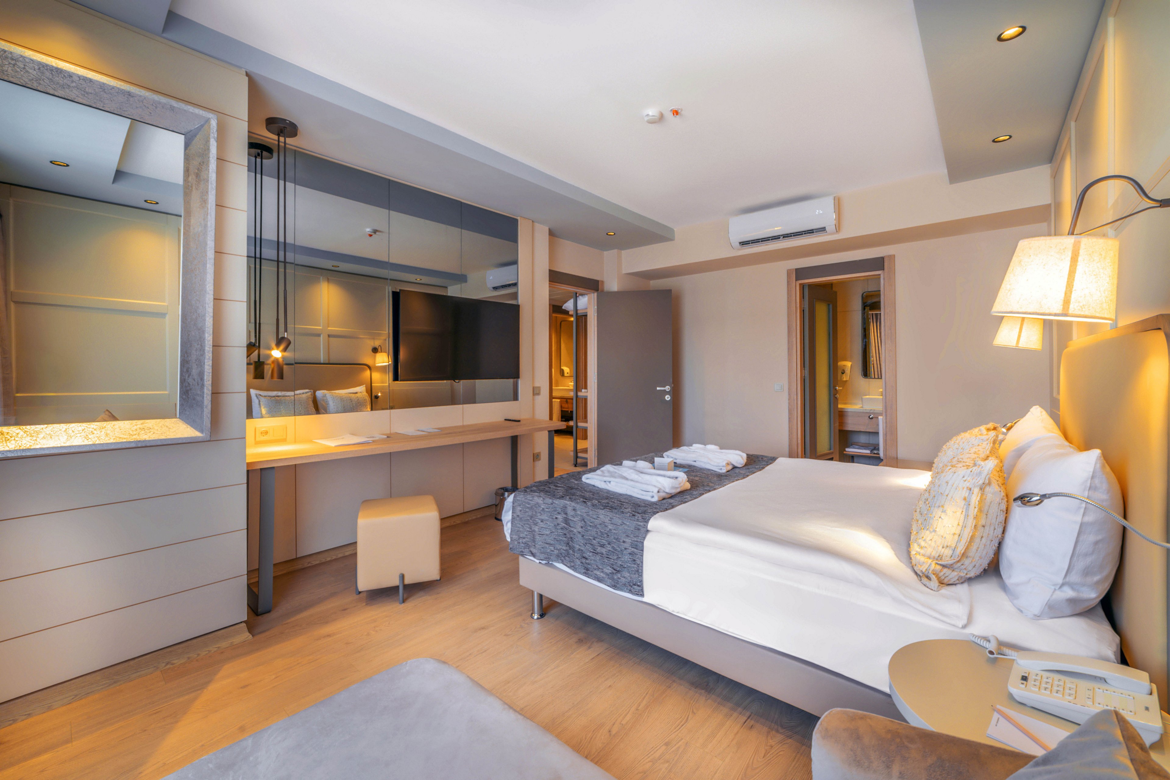 A contemporary hotel bedroom features a neatly made double bed with white linens and decorative pillows. Warm ambient lighting and recessed ceiling lights create a cozy atmosphere, highlighting the neutral beige and gray color palette. A long mirrored vanity with a stool extends along one wall, visually enlarging the space. Towels are folded on the bed, and a bedside table with a telephone completes the inviting, well-appointed room.