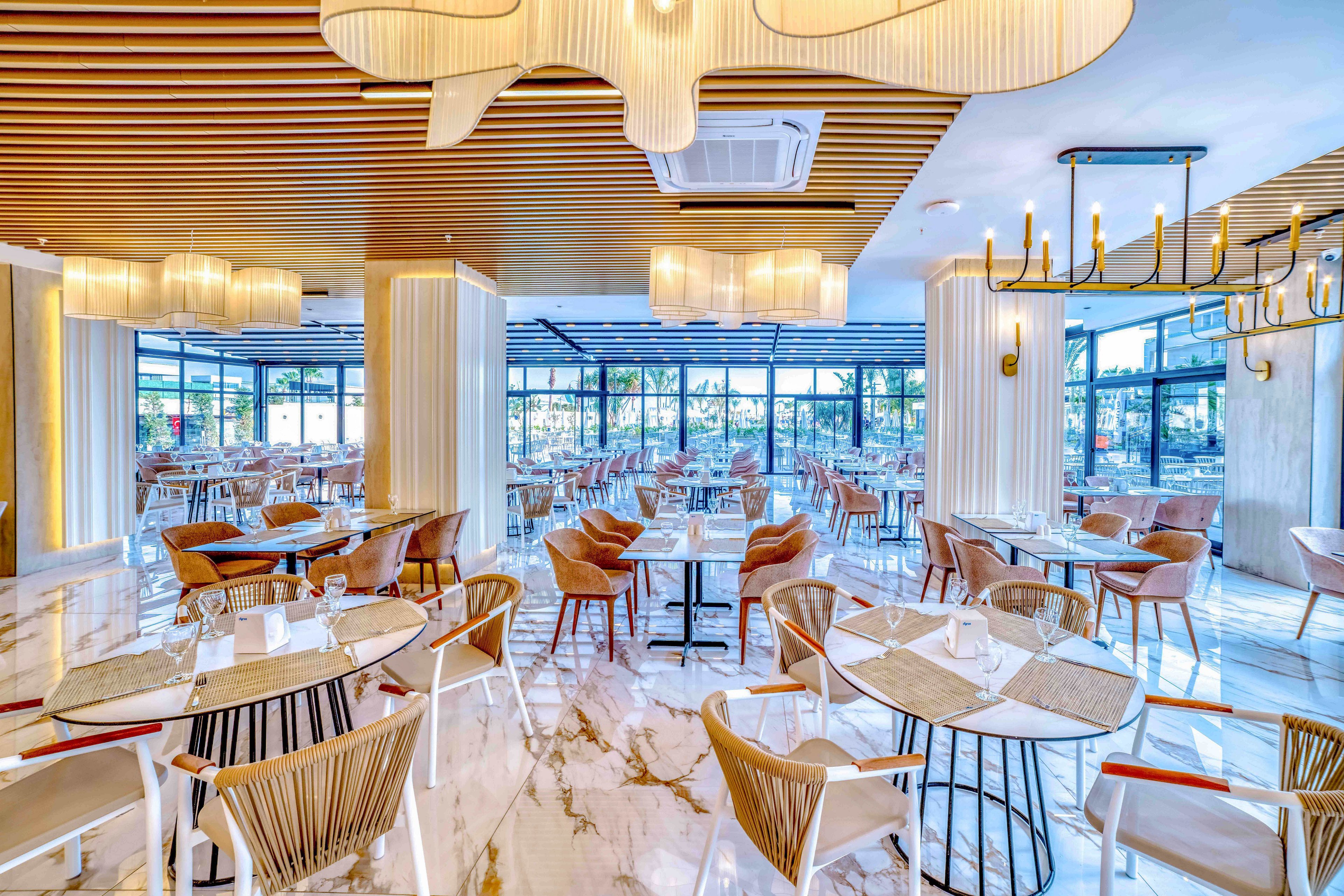 A spacious contemporary restaurant interior features neatly arranged empty tables and chairs on a glossy marble floor. Large windows line the perimeter, allowing natural light to flood the dining area and reveal an outdoor view. Warm wooden accents, sculptural ceiling lights, and elegant chandeliers create a stylish, inviting atmosphere. The setting appears ready for guests, with tableware and placemats already laid out.