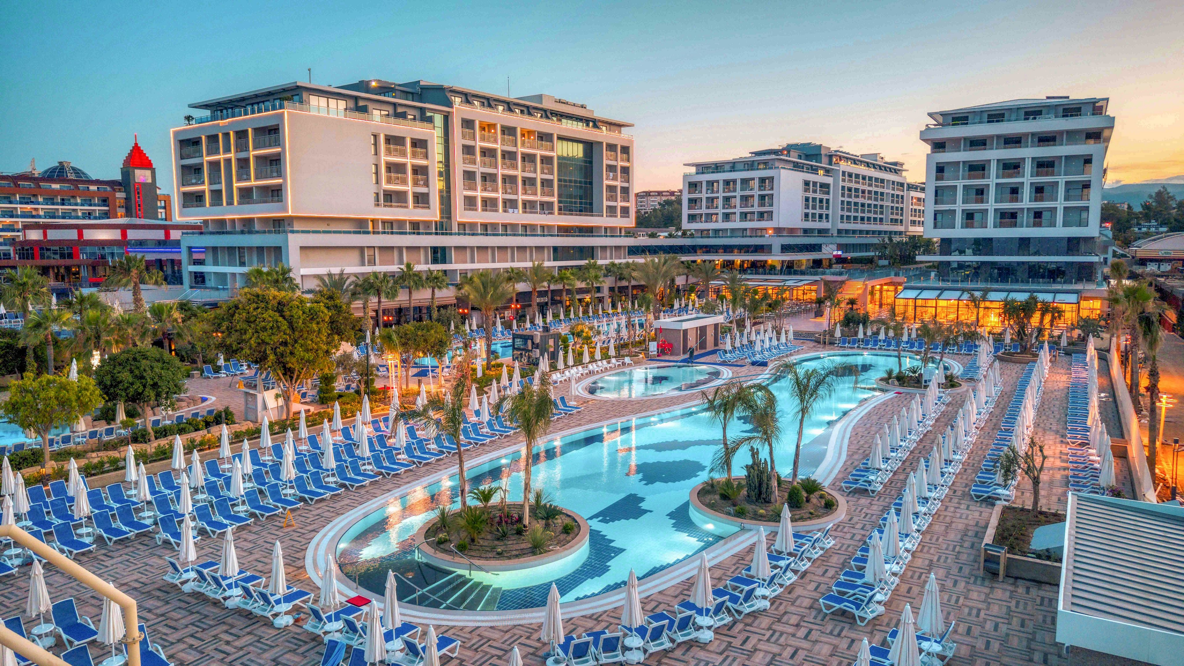A contemporary resort complex features several multi-story hotel buildings surrounding a large, curving outdoor swimming pool. Rows of blue-and-white sun loungers and umbrellas line the poolside terrace, creating a structured, vacation-ready scene. Palm trees and landscaped greenery soften the architecture, while warm evening lights glow from the lower levels. The sky is clear at sunset, adding soft pastel tones to the overall atmosphere.