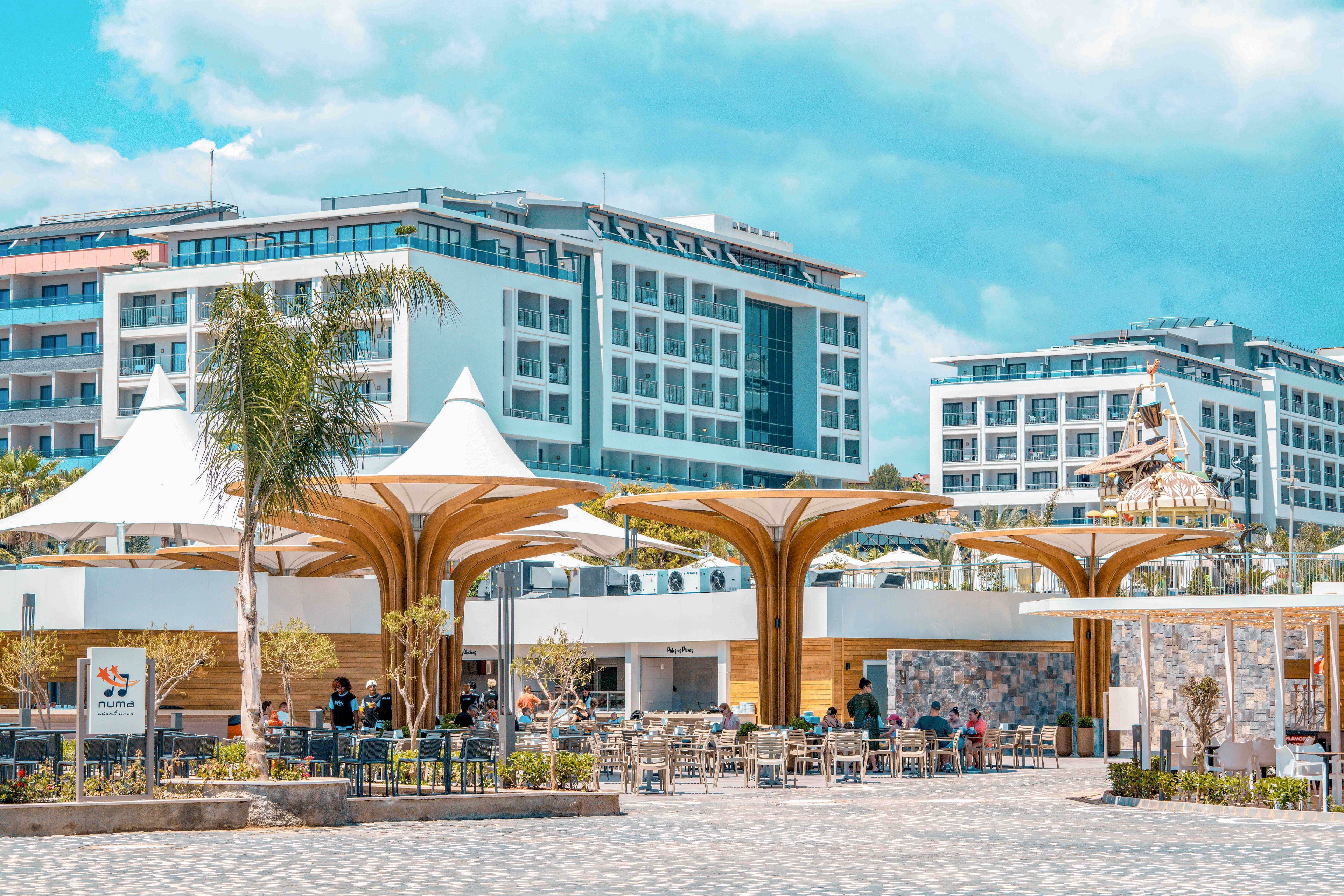 A contemporary urban plaza features striking wooden canopy structures with white fabric tops, creating shaded seating areas for visitors. People sit and walk around an open-air café space, surrounded by palm trees and landscaped planters. In the background, several modern mid-rise apartment or hotel buildings frame the scene under a bright, partly cloudy sky. The overall atmosphere is relaxed and vibrant, suggesting a coastal or resort-style destination.