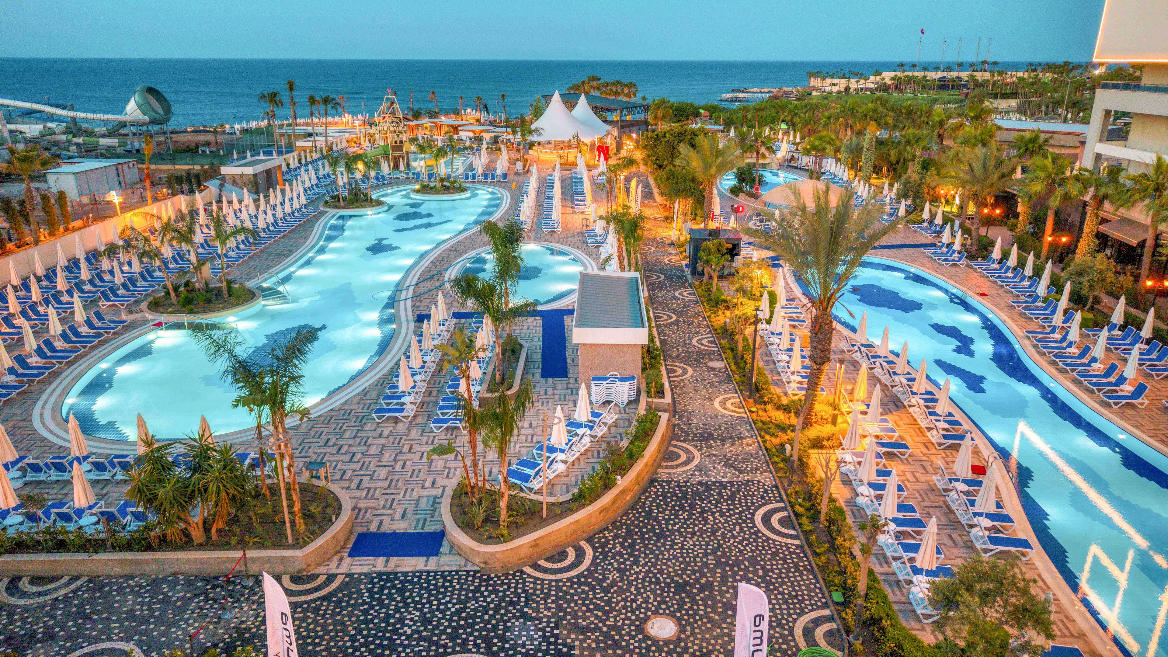 A large luxury resort features multiple winding swimming pools lined with rows of blue and white lounge chairs and umbrellas. Palm trees and landscaped walkways create a tropical atmosphere around the water. The scene overlooks the sea in the background under a soft evening sky. Buildings, cabanas, and recreational areas suggest a popular vacation destination with ample amenities.