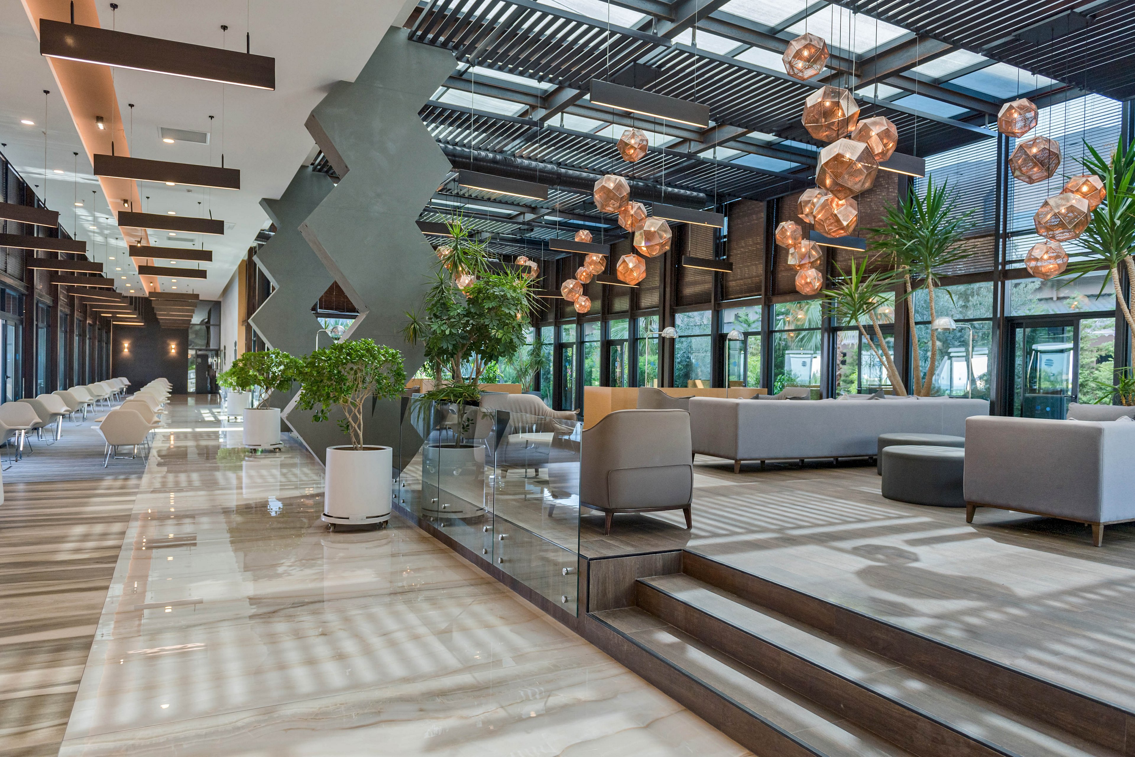 Spacious modern lobby interior featuring sleek sofas and armchairs arranged in multiple seating areas. Large floor-to-ceiling windows line one side, allowing natural light to illuminate the polished floor and glass railings. Decorative pendant lights with warm tones hang from the ceiling, complemented by geometric architectural elements and indoor plants. The overall design feels luxurious, open, and inviting for guests.