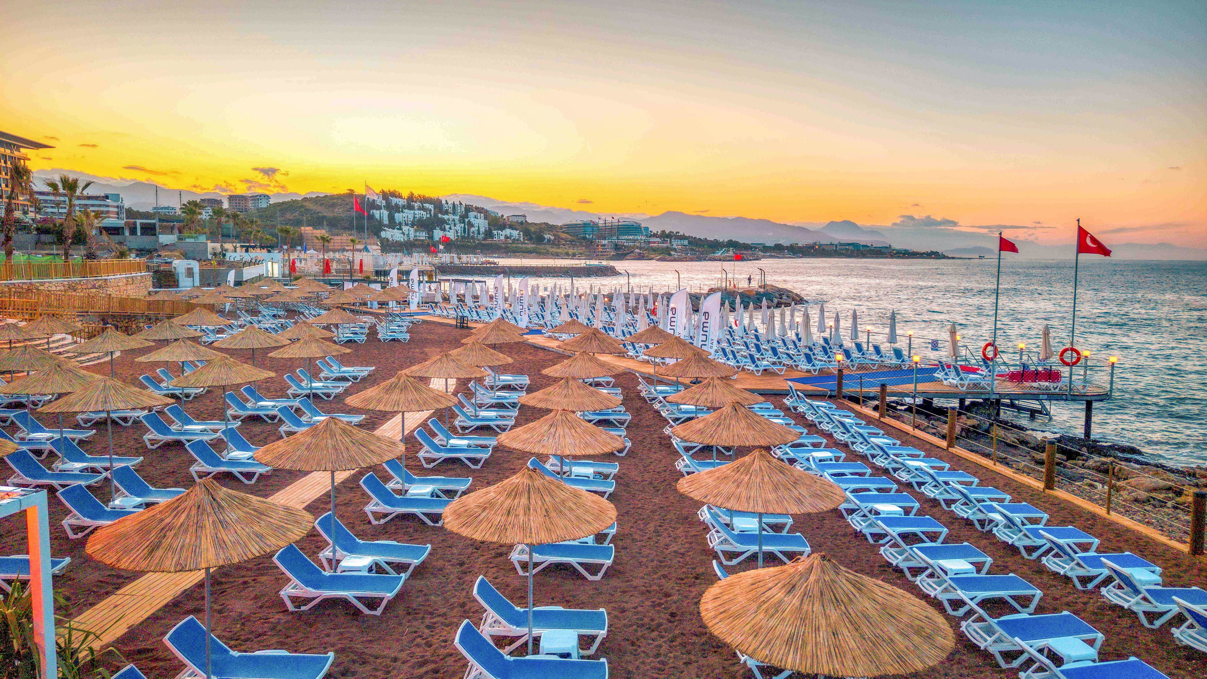 A coastal resort beach is lined with rows of blue sun loungers and straw parasols on reddish sand. The calm sea stretches out toward the horizon under a colorful sunset sky. Red flags and lifebuoys are visible on a wooden pier extending into the water. Buildings and palm trees in the background suggest a popular holiday destination along the shoreline.