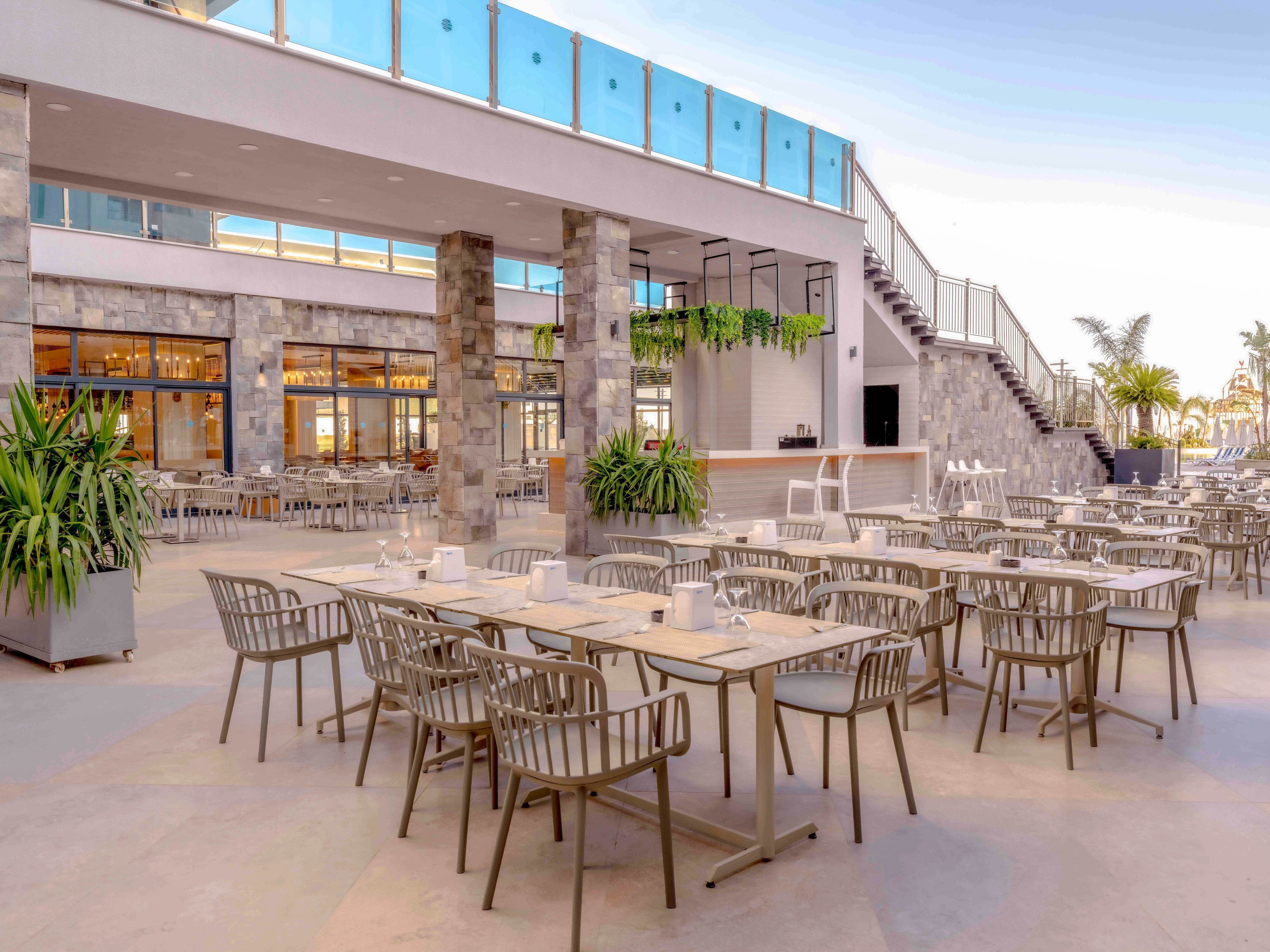 Spacious outdoor dining area with neatly arranged tables and chairs is shown in front of a contemporary building. The setting features a bar counter, staircase to an upper level, and large glass panels along the terrace edge. Green potted plants and hanging foliage add a touch of nature to the clean, modern architecture. Warm evening light creates a relaxed, inviting atmosphere for guests.