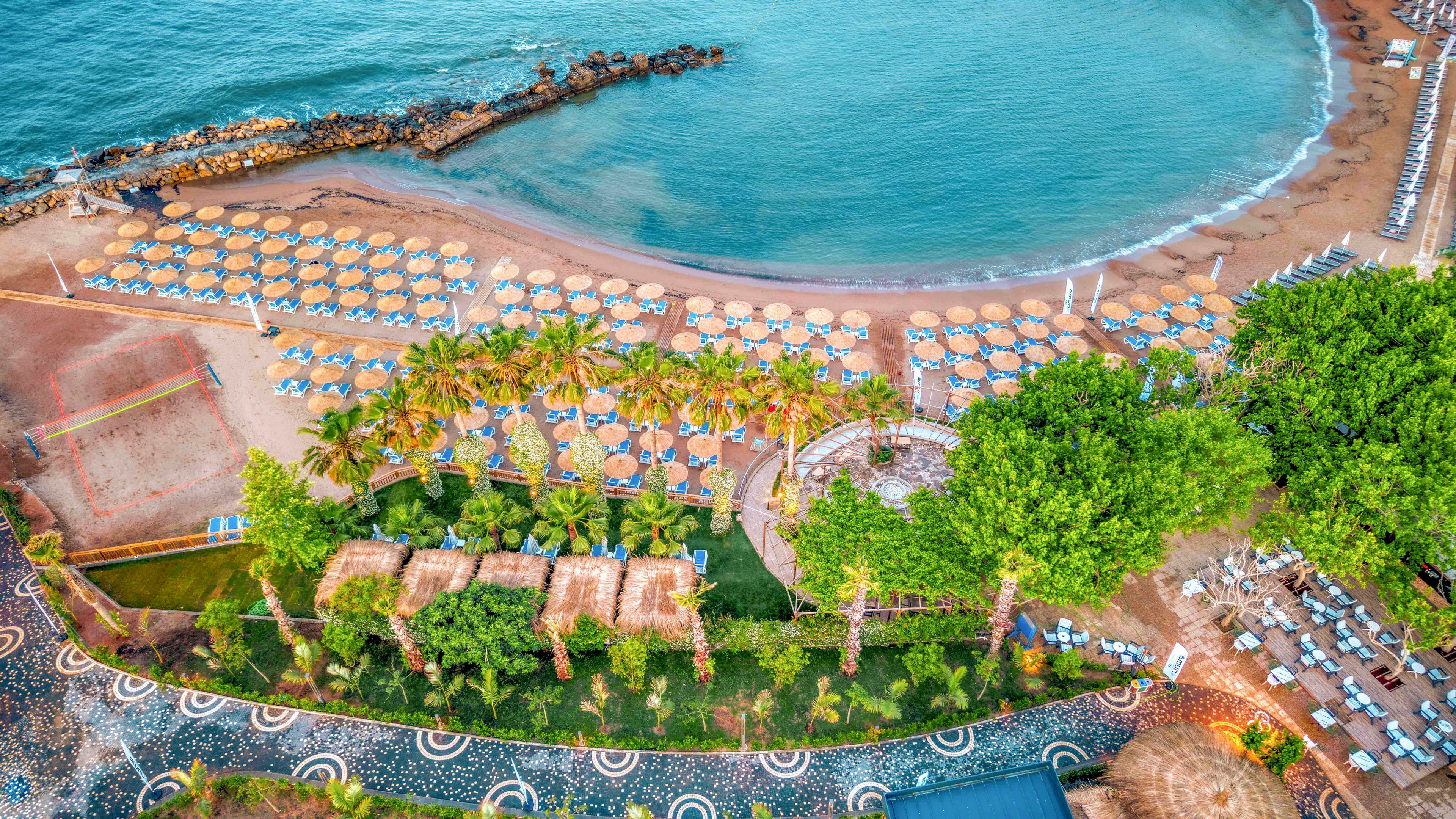 Aerial view of a tropical beachfront resort with rows of sun loungers and umbrellas lining a sandy shore. Calm turquoise water curves around the bay, bordered by a rocky breakwater. Palm trees and lush green gardens separate the beach from the resort pathways and structures. The scene suggests a sunny, relaxing vacation destination with organized beach facilities.