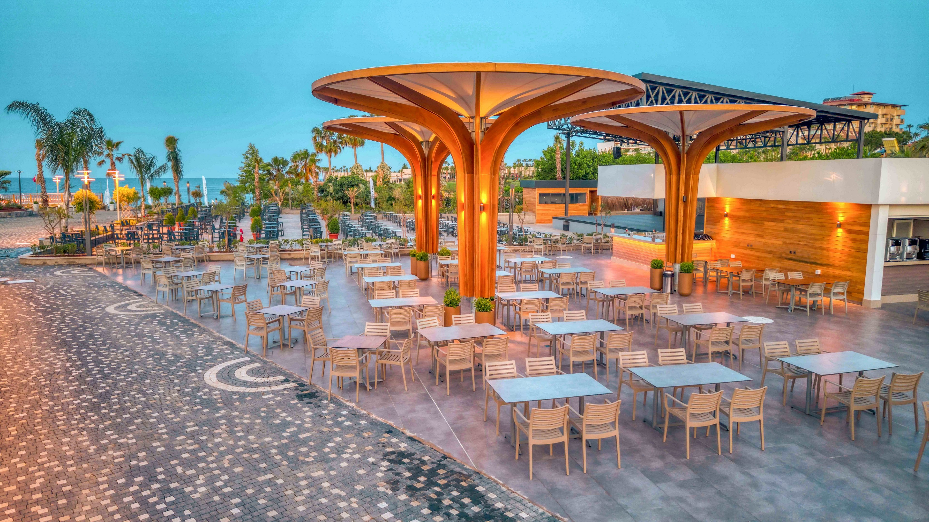 Spacious outdoor dining area with rows of empty tables and chairs is shown under large sculptural wooden canopies. The setting appears to be a modern resort terrace with palm trees and landscaped pathways leading toward the horizon. Warm artificial lighting contrasts with the cool blue evening sky, creating an inviting atmosphere. A bar or service counter is visible to the right, enhancing the impression of a leisure and hospitality environment.