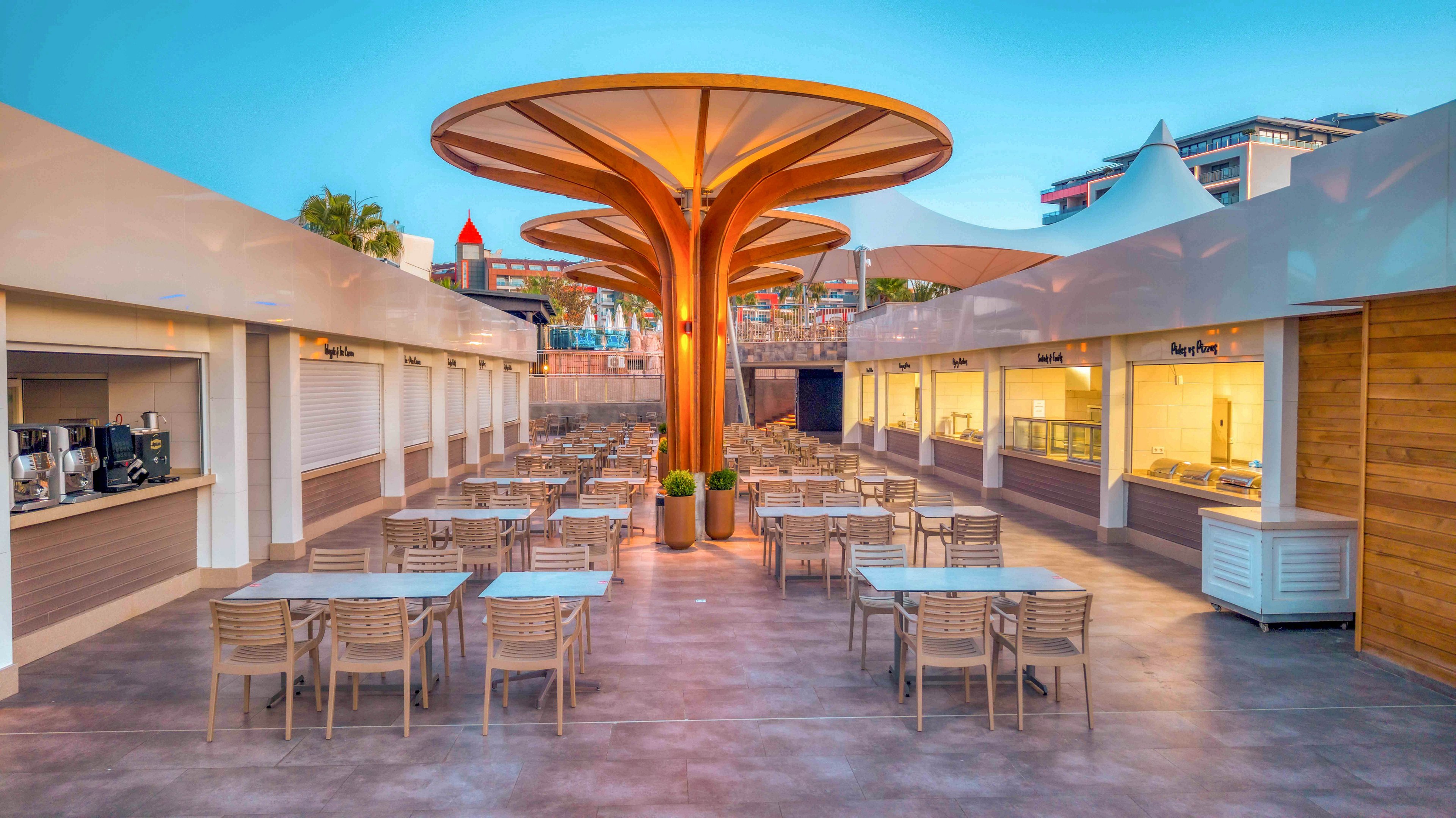 An open-air cafe terrace features rows of empty tables and chairs arranged symmetrically. Large sculptural tree-like columns rise from the center, supporting a canopy structure. Food counters and service areas line both sides under sleek white awnings. The scene is captured at sunset or early evening, with soft blue and warm tones creating a relaxed, contemporary atmosphere.
