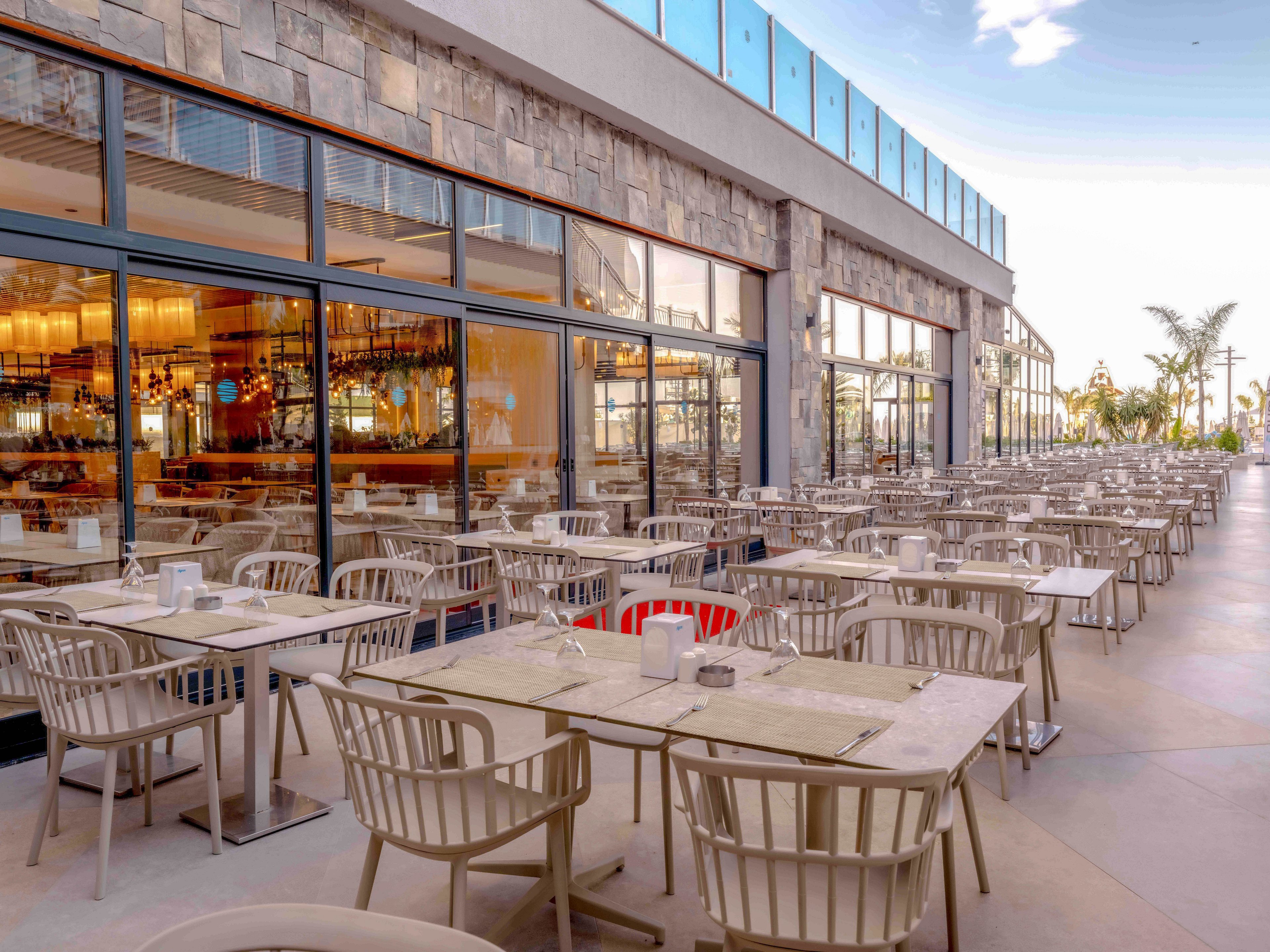 A spacious outdoor restaurant terrace is filled with rows of light-colored tables and chairs. Large glass windows reveal a warm, well-lit interior bar and dining area. The setting appears clean and contemporary, with stone wall cladding and sleek architectural lines. The atmosphere suggests a relaxed, inviting place for al fresco dining in pleasant weather.