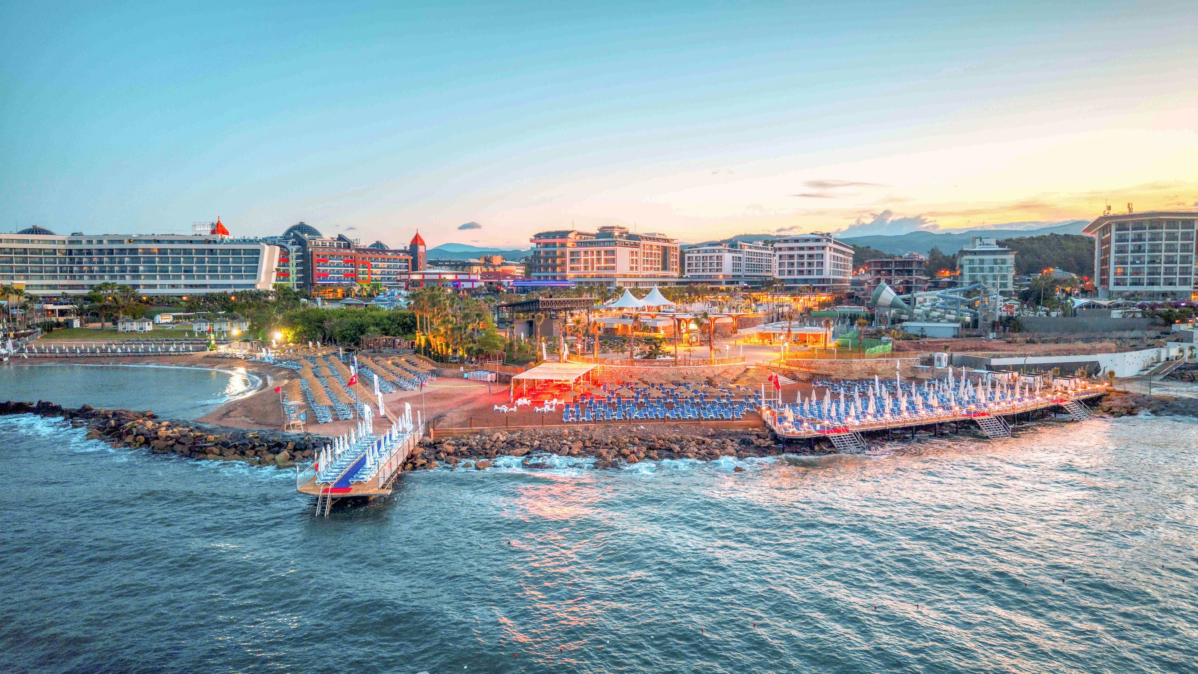 A large seaside resort complex stretches along a rocky coastline at sunset. Multiple hotel buildings face the water, with pools, palm trees, and outdoor seating areas visible. Colorful beach chairs and umbrellas line the shore near a small pier, while warm lights illuminate the central plaza. The calm sea in the foreground reflects the soft evening sky, creating a relaxed vacation atmosphere.