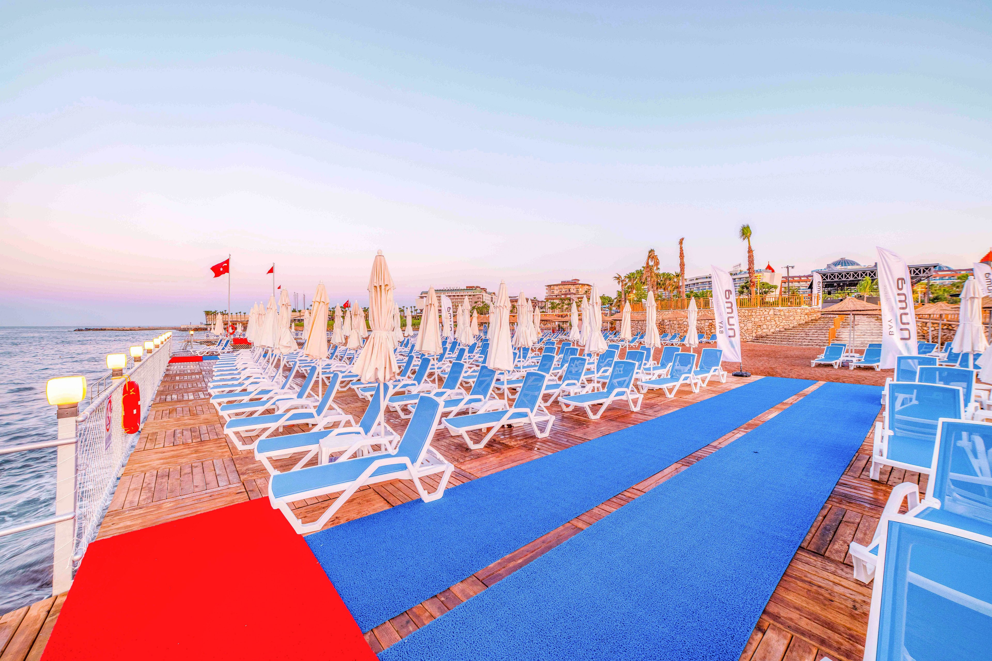 A long wooden pier lined with rows of blue sunbeds and closed white umbrellas stretches out beside a calm sea. Bright red and blue carpets run down the center of the pier, creating a vivid contrast against the warm evening sky. Resort buildings and palm trees are visible in the background, suggesting a coastal holiday destination. The scene feels peaceful and quiet, with no people present and soft pastel colors in the sky.
