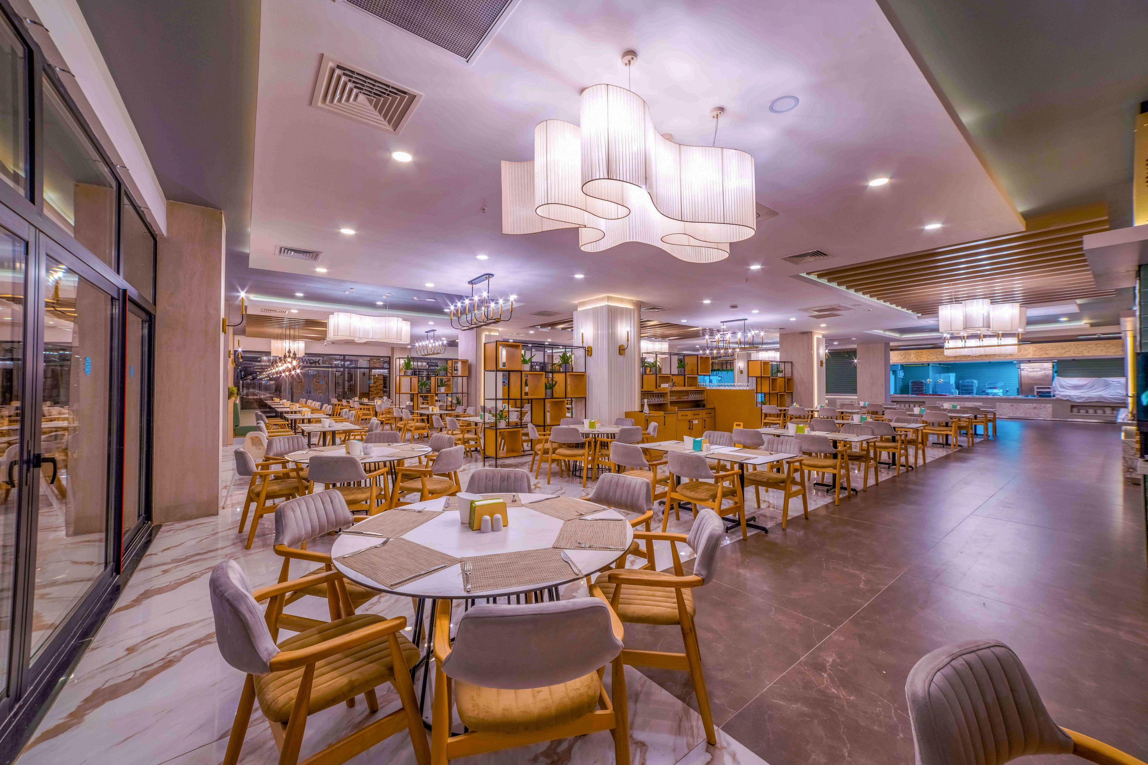 Spacious contemporary restaurant interior with neatly arranged round tables and wooden chairs. The setting appears to be an empty dining area, featuring soft ambient lighting and decorative ceiling fixtures. Large pendant lamps and chandeliers create a warm, inviting atmosphere, complemented by neutral tones and clean lines. Glass walls, tiled flooring, and an open layout emphasize a modern, upscale design suitable for hospitality or events.