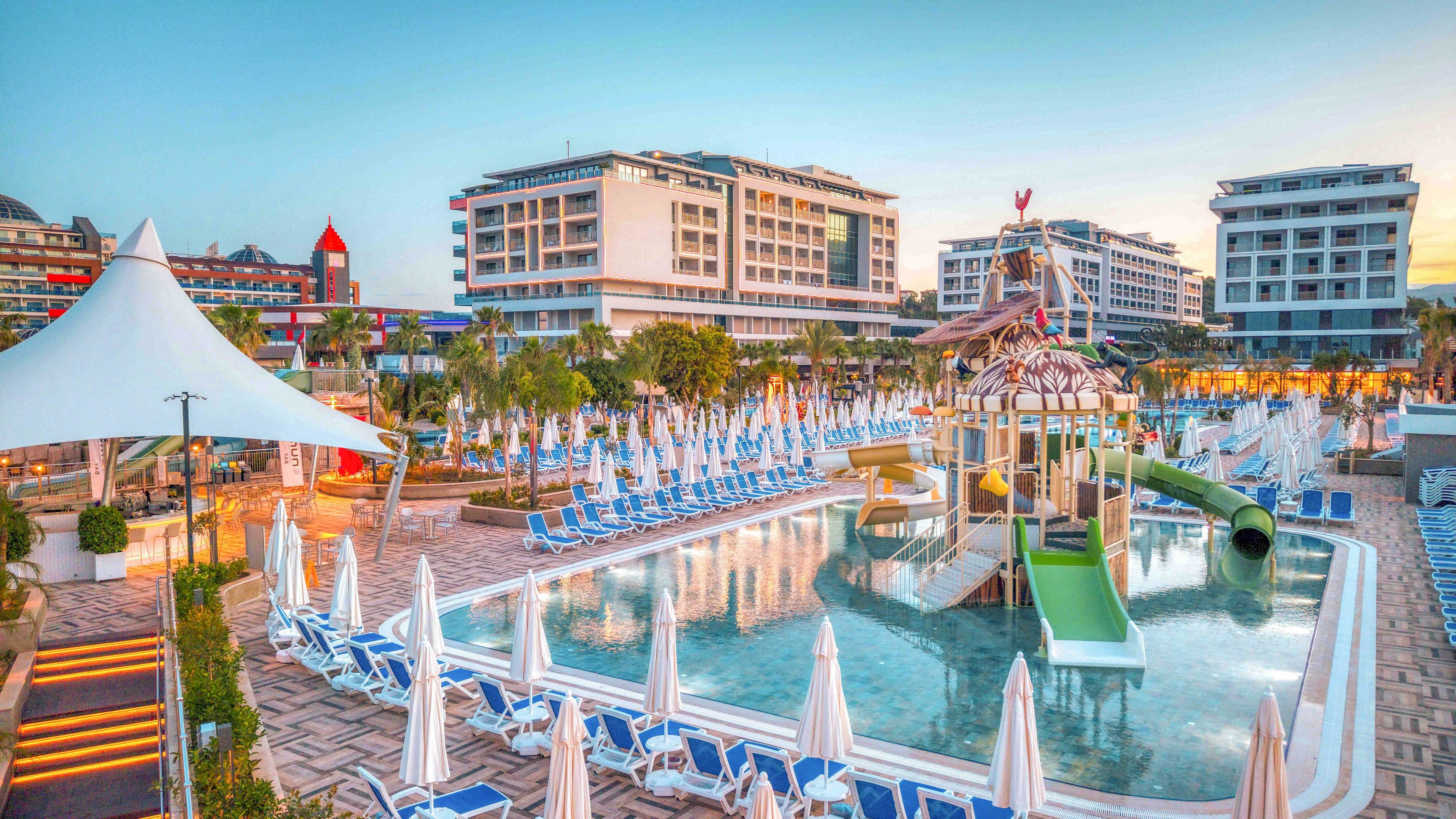 A large resort hotel complex features an expansive outdoor swimming pool with a colorful water park structure and slides. Numerous blue sun loungers and white umbrellas line the poolside, creating a well-organized relaxation area. Contemporary multi-story hotel buildings surround the pool, with palm trees and landscaped walkways adding a tropical vacation feel. The scene is captured at sunset or early evening, with soft warm light enhancing the tranquil holiday atmosphere.