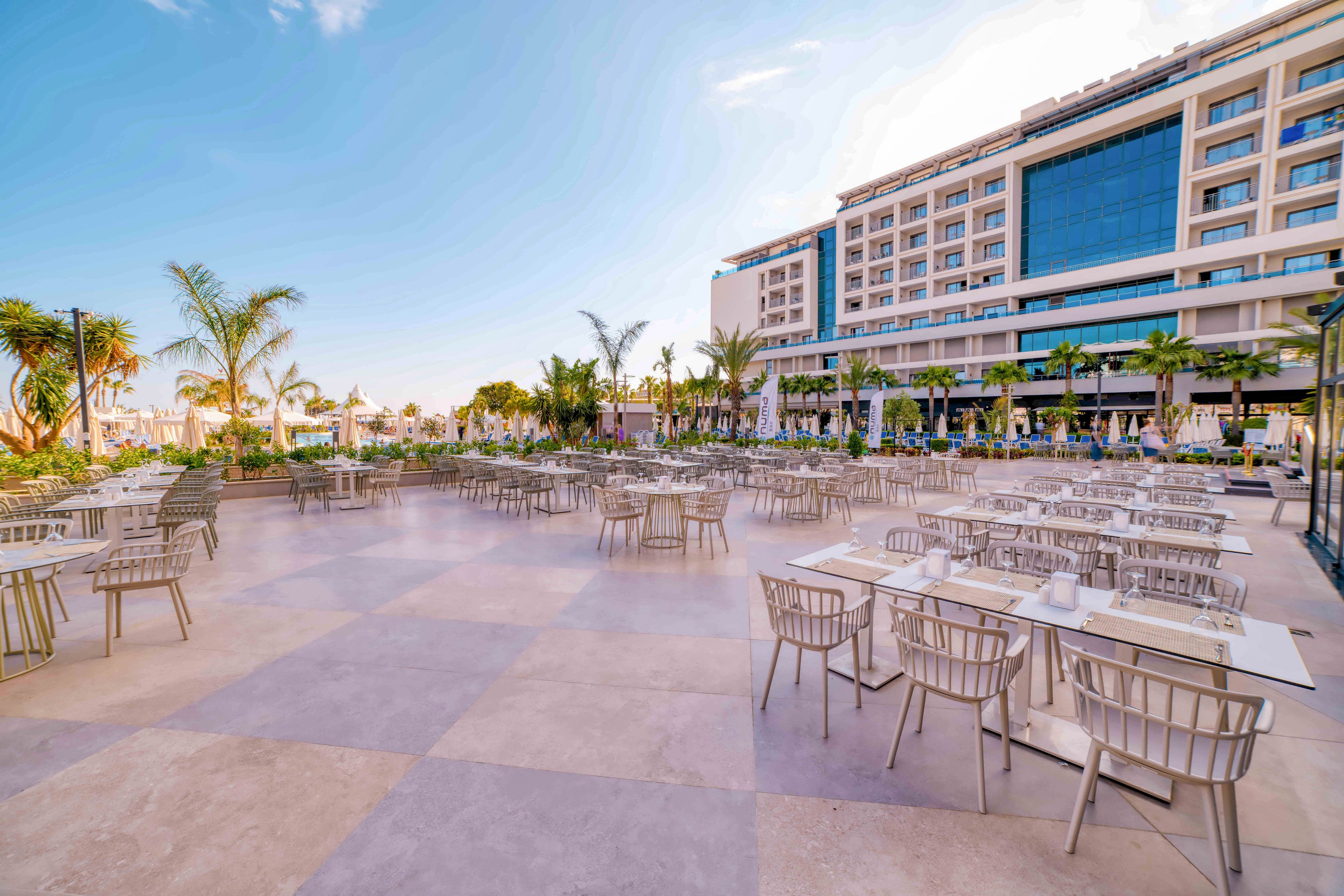 Spacious outdoor dining terrace with numerous tables and chairs is set in front of a modern multi-story resort hotel. Palm trees and landscaped greenery frame the open plaza, creating a relaxed vacation atmosphere. The sky is clear with warm light, highlighting the contemporary architecture and inviting seating. No people are present, emphasizing the clean, orderly setup of the restaurant area.