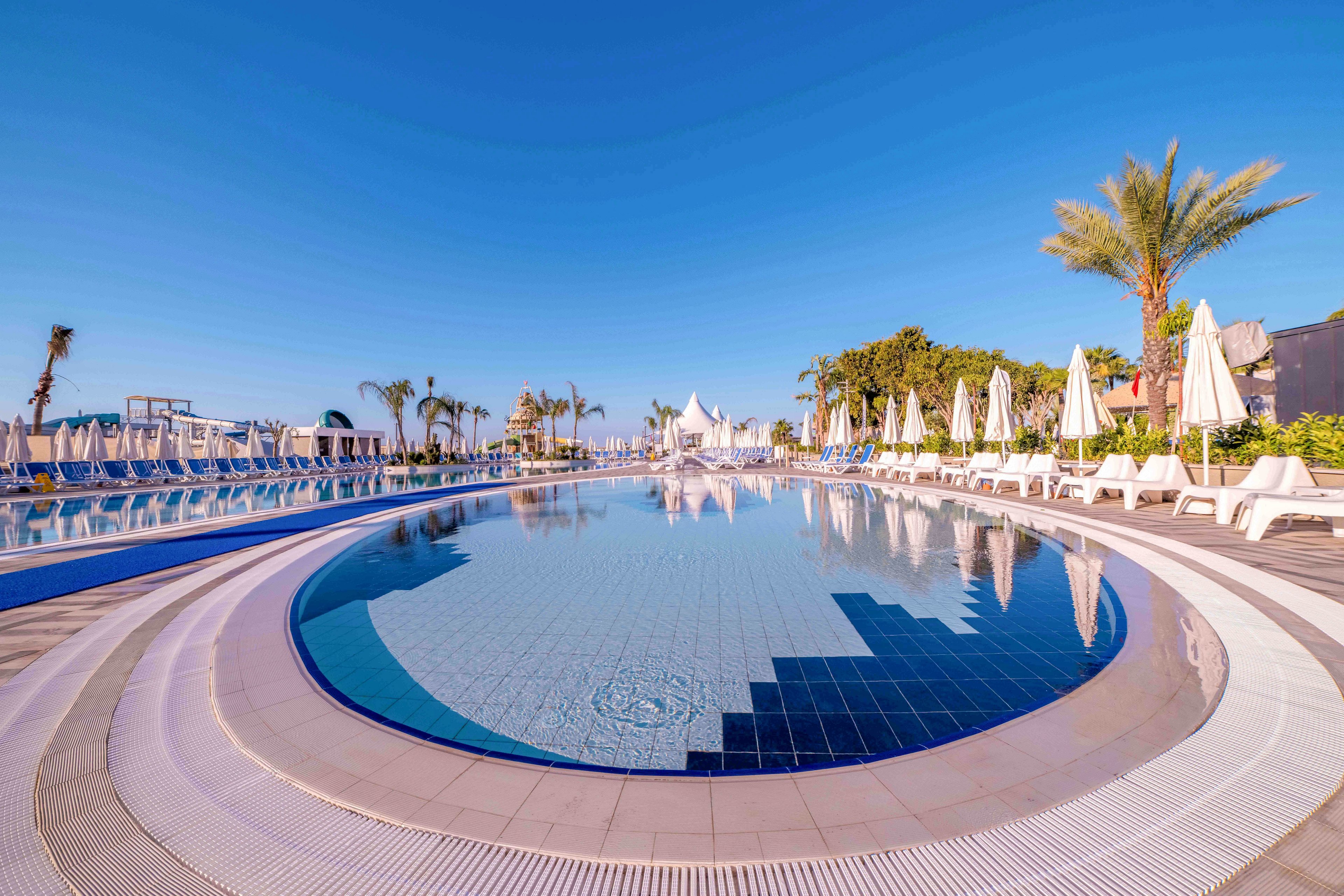 A large circular swimming pool is featured in an outdoor resort setting under a clear blue sky. White sun loungers and umbrellas line both sides of the pool, suggesting a relaxing vacation atmosphere. Palm trees and landscaped greenery frame the scene, adding a tropical feel. The calm water reflects the surrounding structures and bright daylight, emphasizing a peaceful and inviting mood.