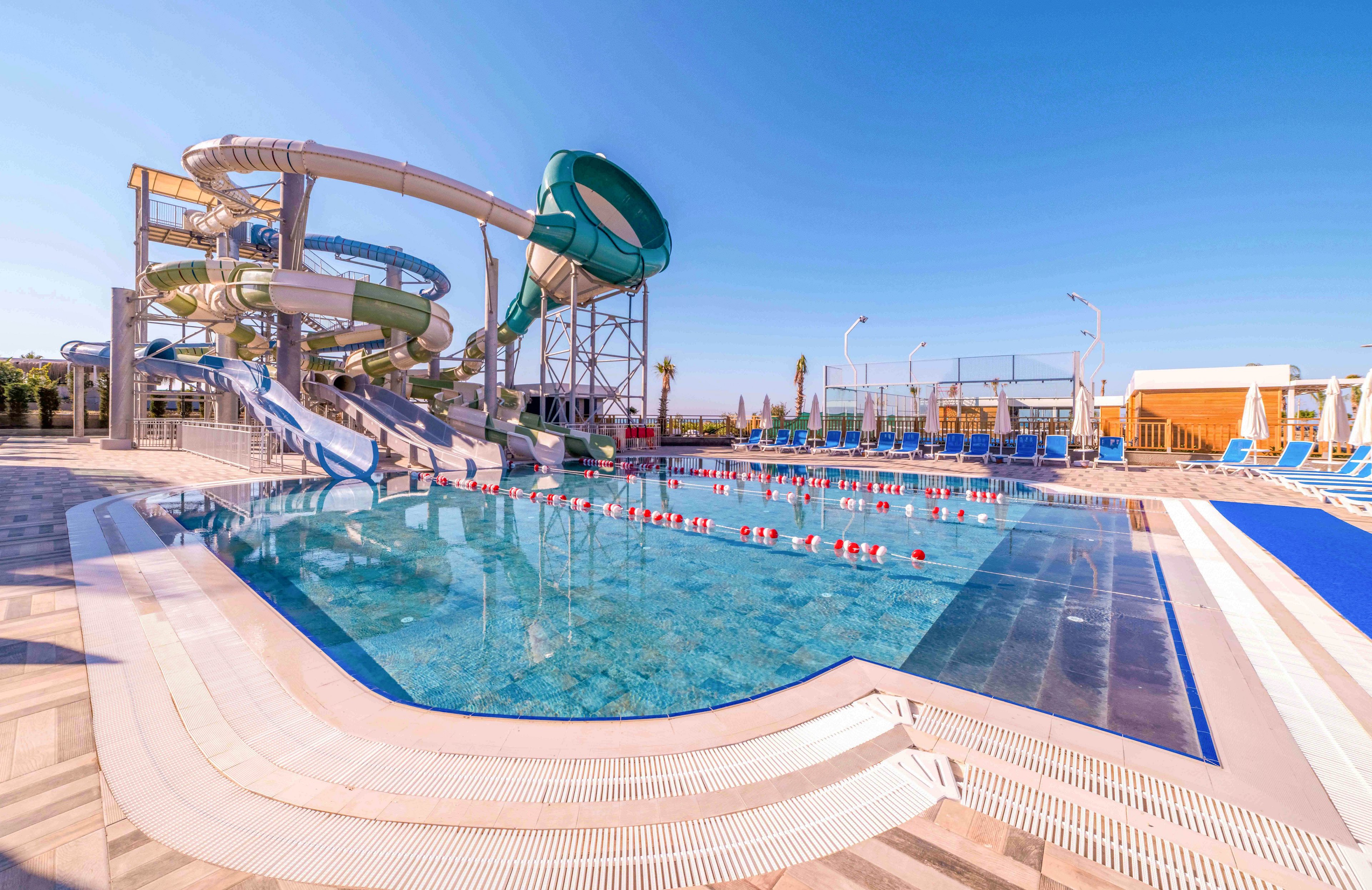 An outdoor swimming pool is shown in front of a large water slide structure under a clear blue sky. Multiple twisting slides descend into the pool area, with lane dividers and floating markers visible in the water. Sun loungers and umbrellas line the deck, suggesting a leisure and vacation setting. The bright sunlight and clean, modern design create a fun and inviting atmosphere.